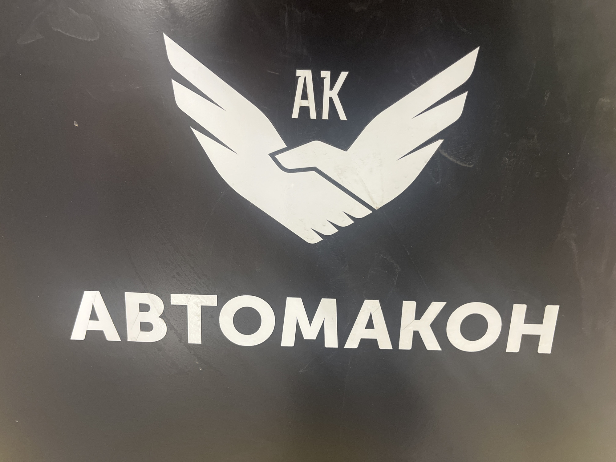 The image displays a white logo and text against a dark, possibly black, background, likely part of a sign or panel in Lupanovo, Russia. The central element of the logo is a stylized graphic of two hands clasped in a handshake, forming a shape reminiscent of outstretched wings. Above this graphic, the letters "AK" are clearly visible in white capital script. Below the logo, the Cyrillic word "АВТОМАКОН" is spelled out in large, bold white capital letters. The dark background surface exhibits minor scuffs, dust, and smudges, indicating some wear or use. No specific time of day, weather, or activity is depicted beyond the static display of the logo.