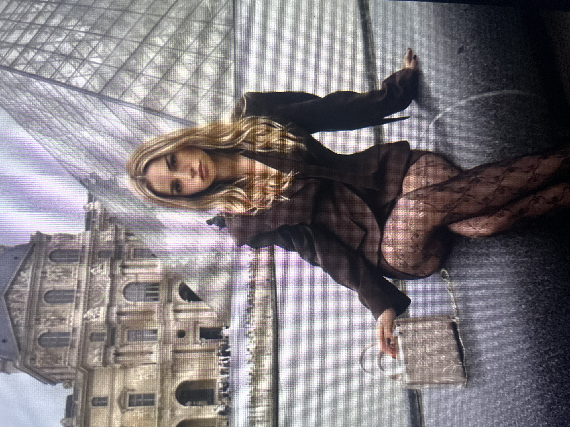 A young woman with long blonde hair is sitting on a dark gray stone bench in front of a modern glass pyramid, with a classical building in the background. She is wearing a dark brown blazer with padded shoulders, a matching skirt, and black patterned stockings. She is holding a small silver handbag with a chain strap. Her left hand rests on the handbag, and her right hand is extended towards it. The background shows the Louvre Museum in Paris, France. The image appears to be a photograph taken on a screen or monitor, indicated by the pixelated quality. There are no visual cues to suggest this location is in Kołobrzeg, Poland.