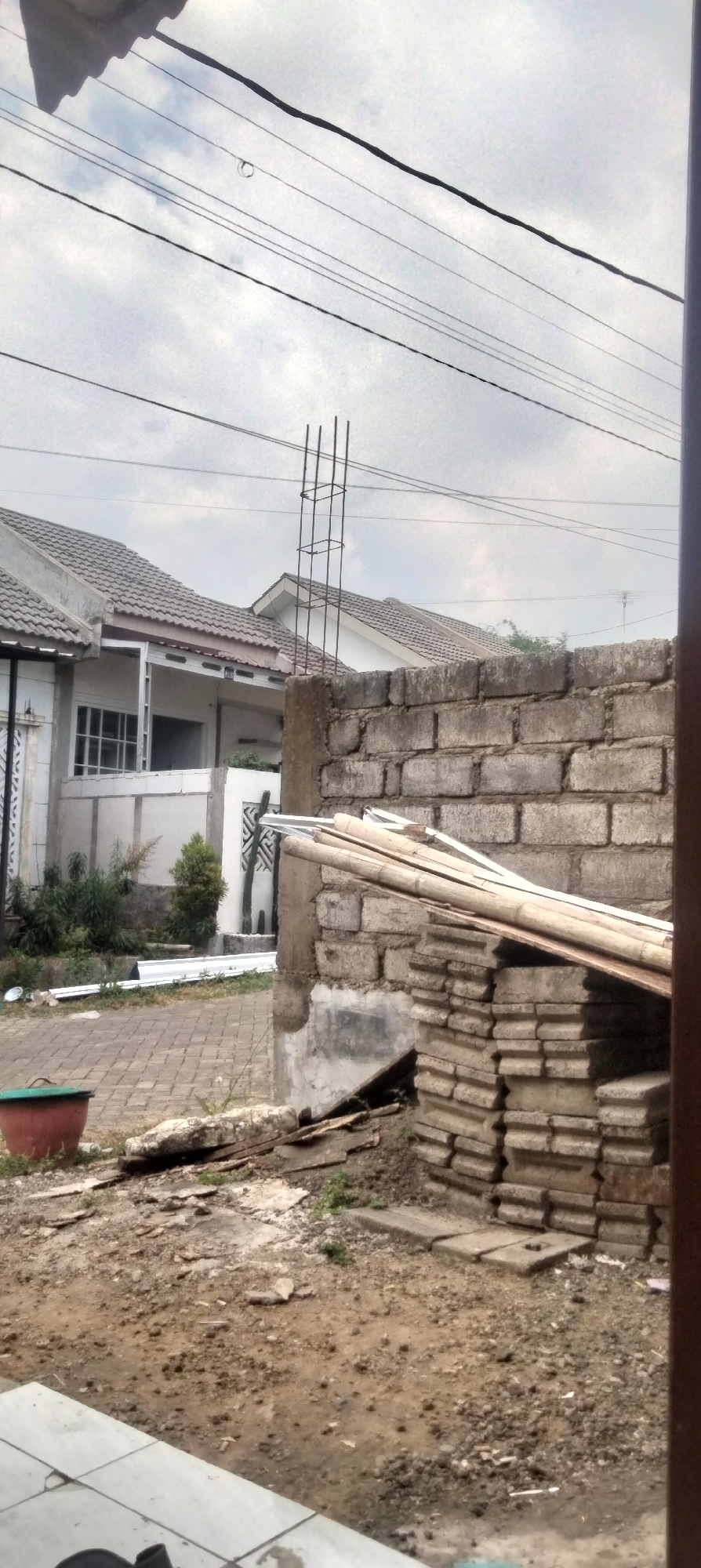 The image depicts an outdoor scene, likely in Antirogo, Indonesia, characterized by construction materials and residential structures. In the foreground, the ground is uneven and consists of dirt and debris, with a reddish-brown pot visible on the left. A brick wall, partially constructed or in disrepair, dominates the middle right of the frame, with stacked concrete blocks and bamboo poles leaning against it. Behind the wall, a metal rebar structure, resembling part of a pillar, extends upwards against a cloudy sky. Further in the background, two adjacent houses with grey tiled roofs are visible. One house has a white facade with visible window frames, while the other is mostly obscured. Several utility wires crisscross the sky above. The environment suggests an ongoing construction or renovation project within a residential area.