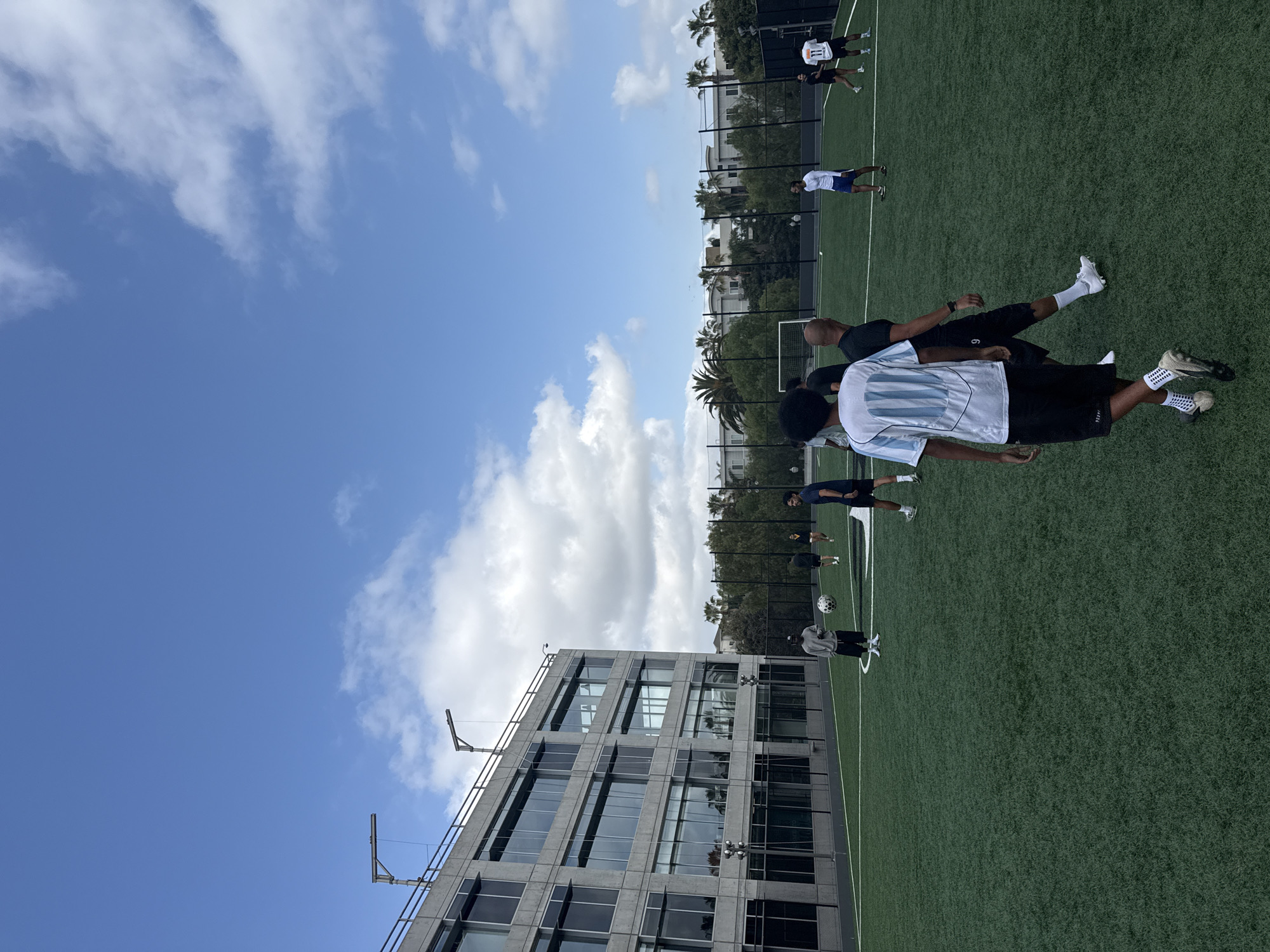 The image depicts a group of people playing soccer on an artificial turf field adjacent to a modern, multi-story building with a grid-like facade in Los Angeles, United States of America. The sky is partly cloudy. The building appears to be under construction, with scaffolding visible on its exterior. The soccer field is enclosed by a black fence, with trees and other buildings visible beyond. Several players are wearing soccer uniforms, while others are in casual attire. Players are actively engaged in soccer-related activities.
