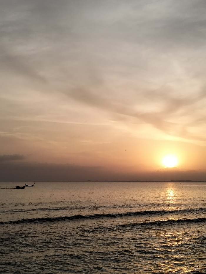 A serene sunrise over the ocean paints the sky in hues of orange and gray. The sun, a radiant orb, casts a glimmering reflection across the rippling water. Small waves gently lap the shore. Two boats are visible in the distance, silhouetted against the horizon. The scene exudes tranquility and peace.