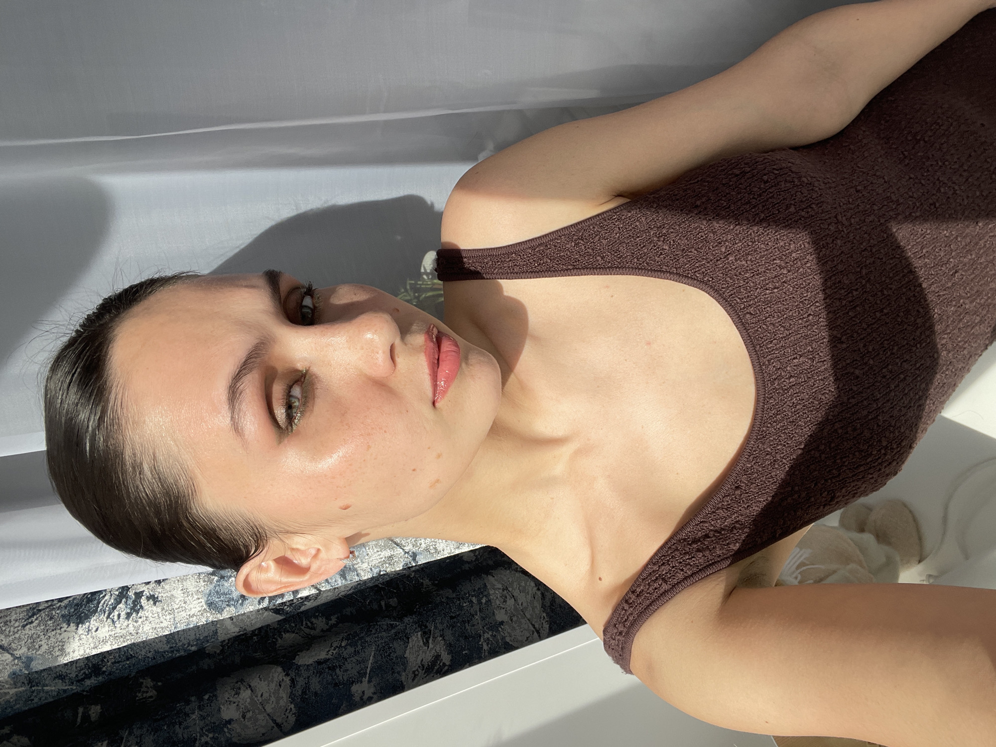 A woman is lying down, facing the camera. She has light skin, dark hair pulled back, and light green eyes. She is wearing a brown tank top. The background appears to be a window with a white curtain partially obscuring the view. Below the window is a patterned surface, possibly a windowsill or floor. There is no indication of the location being Izhevsk, Russia.