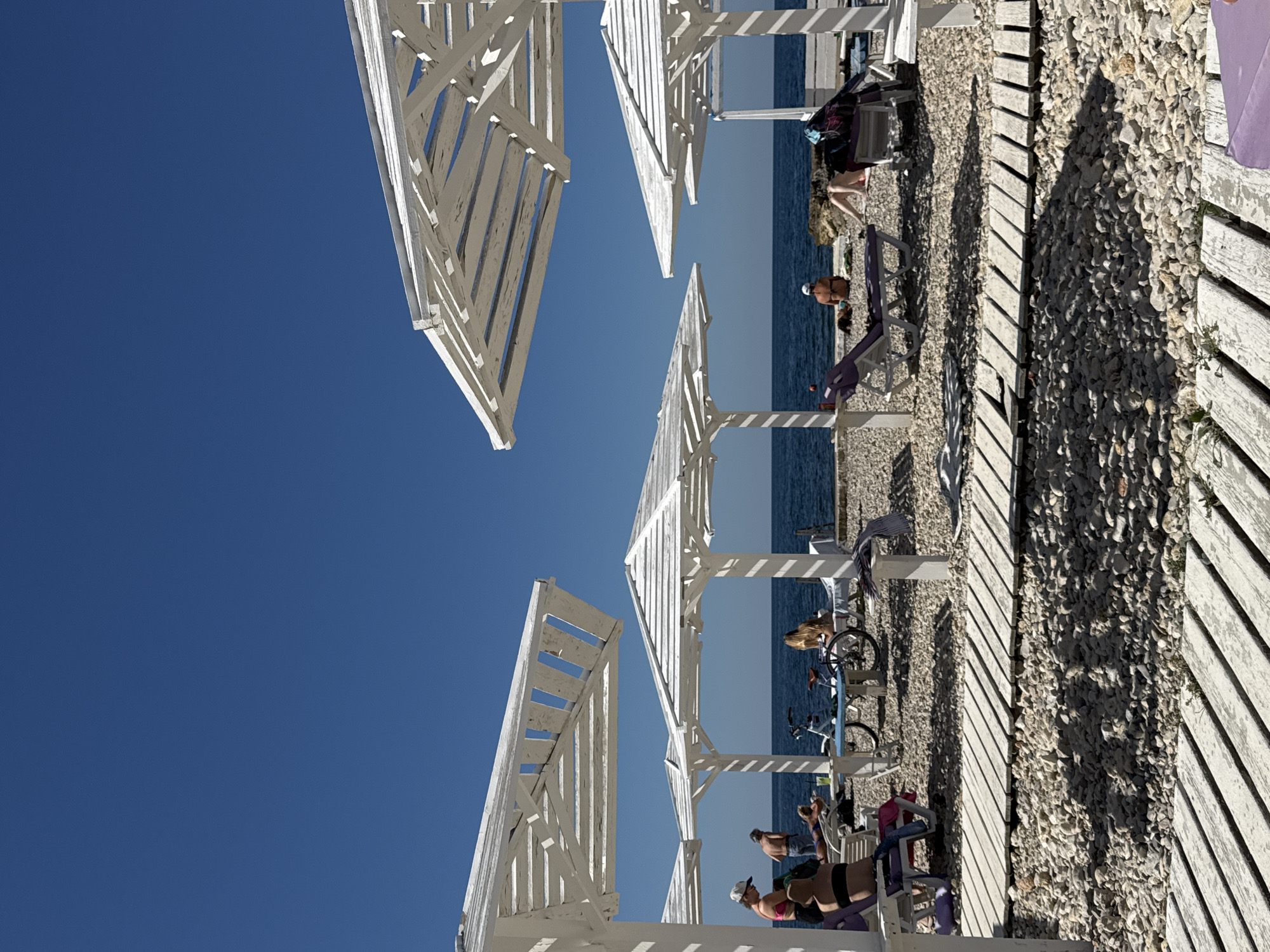The image depicts a sunny day at a pebble beach in Sevastopol, Ukraine. The scene is dominated by several white wooden beach umbrellas, casting shadows on the ground. In the foreground, two wooden walkways run horizontally across the frame, separated by a strip of white pebbles.
Several people are visible on the beach, relaxing on lounge chairs under the umbrellas. One person in the foreground, partially visible on the left, is wearing a black bikini top and has dark hair. Further back, another person is seen lying on a lounge chair. There are also bicycles and other beach furniture scattered around. The sea is calm and blue, with a clear, deep blue sky overhead. The overall impression is one of a peaceful and pleasant day at the beach.