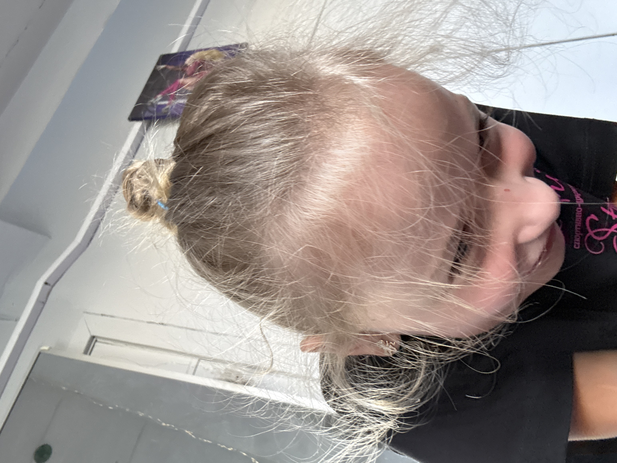 This is a close-up, low-angle, rotated shot of a young person's head and shoulders. The subject has fine, light-colored hair that is messy and stands out in wisps around their head. A small bun is visible at the back of their head, secured with a blue hair tie. Their skin appears fair. The person is wearing a black t-shirt with some pink text and a design on the shoulder area. The visible text appears to be in Cyrillic script, possibly relating to a logo or branding.
The background is out of focus but shows elements of an indoor setting. There are white walls and possibly a shelf or frame with some blurred imagery on it. The lighting suggests it is daytime, with natural light contributing to the scene. The overall impression is casual, possibly a candid shot taken indoors. The subject's expression is partially visible, with one eye closed and the mouth slightly open, suggesting a relaxed or playful mood. The context of Dnipro, Ukraine, does not provide specific visual cues within the image itself.