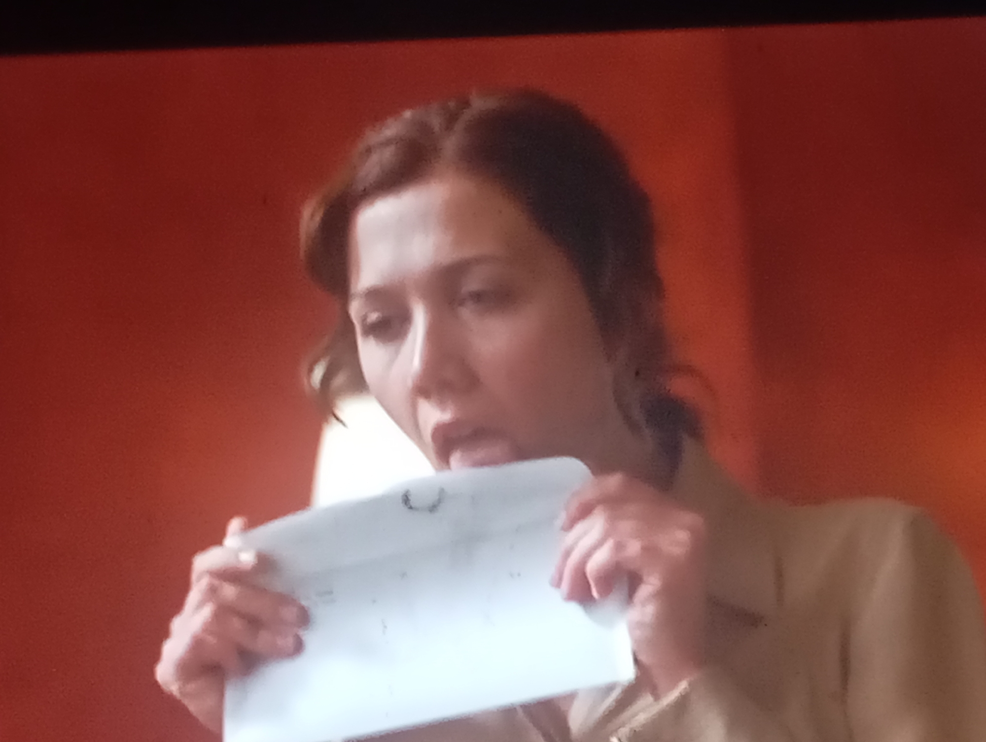 The media file shows a close-up of a woman with brown hair, wearing a tan jacket. She is holding a white envelope in her hands and appears to be licking the flap to seal it. Her facial expression is neutral, and her eyes are looking down at the envelope. The background is a solid, warm orange color, possibly a wall. The lighting is somewhat dim, suggesting an indoor setting, perhaps in the evening or on an overcast day. There are no discernible weather cues, location-specific details beyond the given context of Sofiivska Borshchahivka, Ukraine, or any visible text. The focus is entirely on the woman and her action of sealing the envelope.