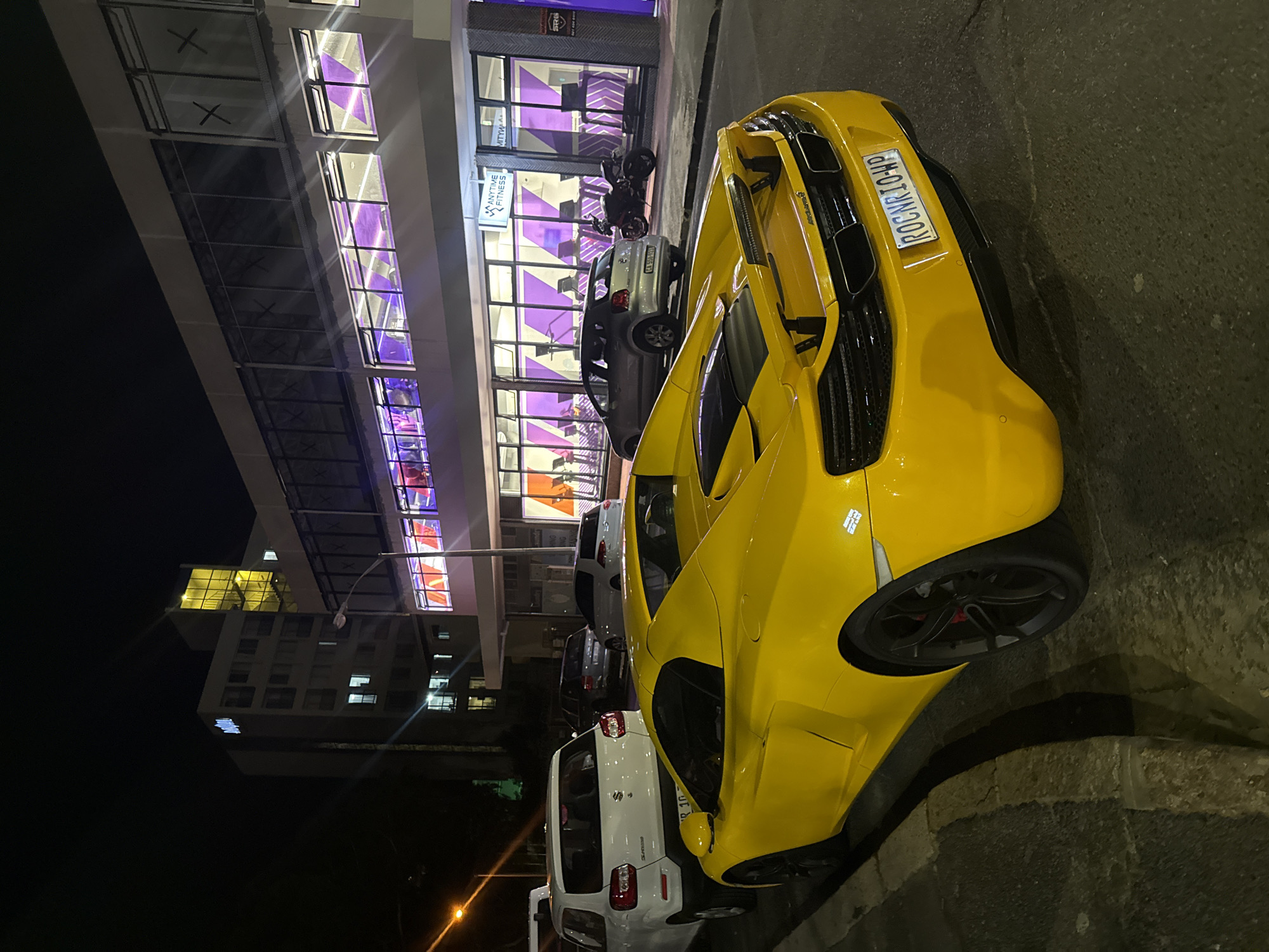 At night, a yellow sports car with the license plate "ROCNK10-HP" is parked on a street in Cape Town, South Africa. The car is alongside a sidewalk with several other parked vehicles, including a white SUV. A building with illuminated purple signs, which may be a gym, is in the background. A multi-story building is also visible in the distance. The lighting is mixed, with streetlights and building lights illuminating the scene.