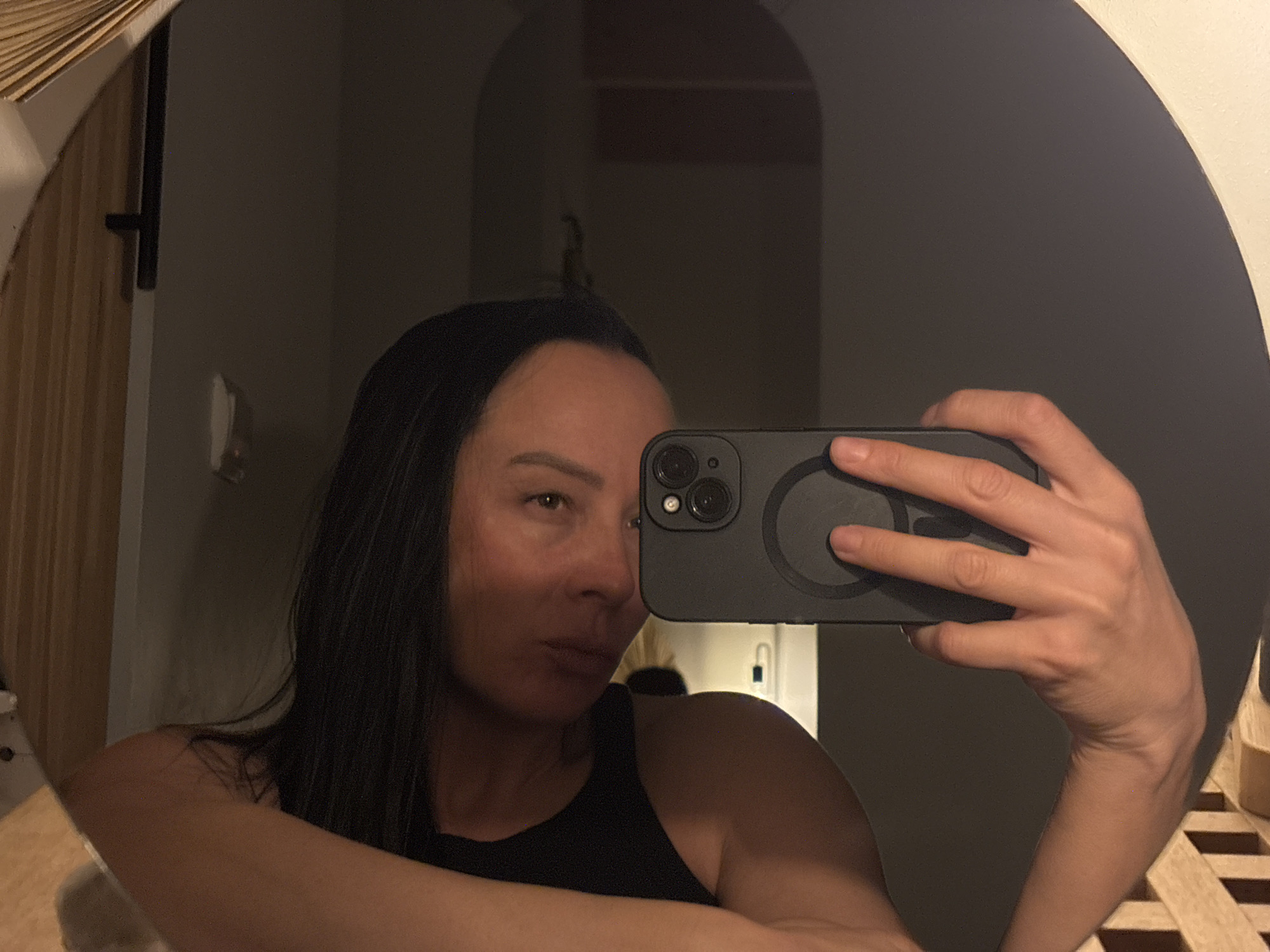 A person is taking a selfie in a mirror. The person has dark hair and is wearing a black tank top. The phone being used is black and has a circular magnet on the back. The mirror appears to be round and has a decorative frame. In the background, there is a glimpse of a room with a partially visible doorway or archway. No information suggests the image was taken in Chaikovsky, Russia.