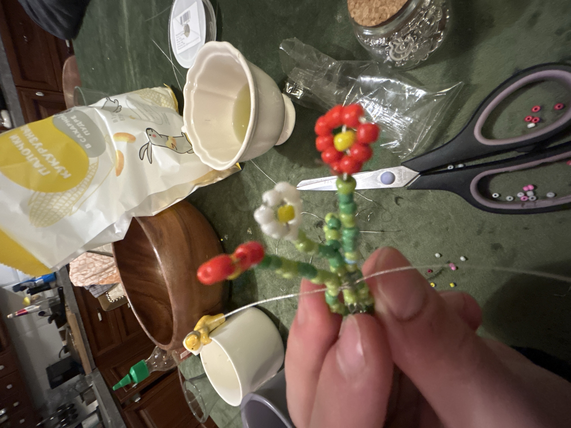 A medium-angle, eye-level shot shows a close-up view of a bead flower being held in a person's hand. The flower has a red beaded top with a yellow center, a white beaded flower with a yellow center, and a red bud. The stem and leaves are made of green beads.
In the background, various objects are scattered on a dark green surface, including a bag of "ПАЛОЧКИ КУКУРУЗНЫЕ" (corn sticks), a small white bowl with a liquid inside, a wooden bowl, and a small white cup with a figurine attached. There's also a pair of black-handled scissors, a jar filled with small metal charms, and a spool of clear thread.
Scattered around the surface are a few loose beads of different colors. The background includes hints of a kitchen setting with wooden cabinets. The overall lighting is moderate, providing adequate illumination for the objects in view.