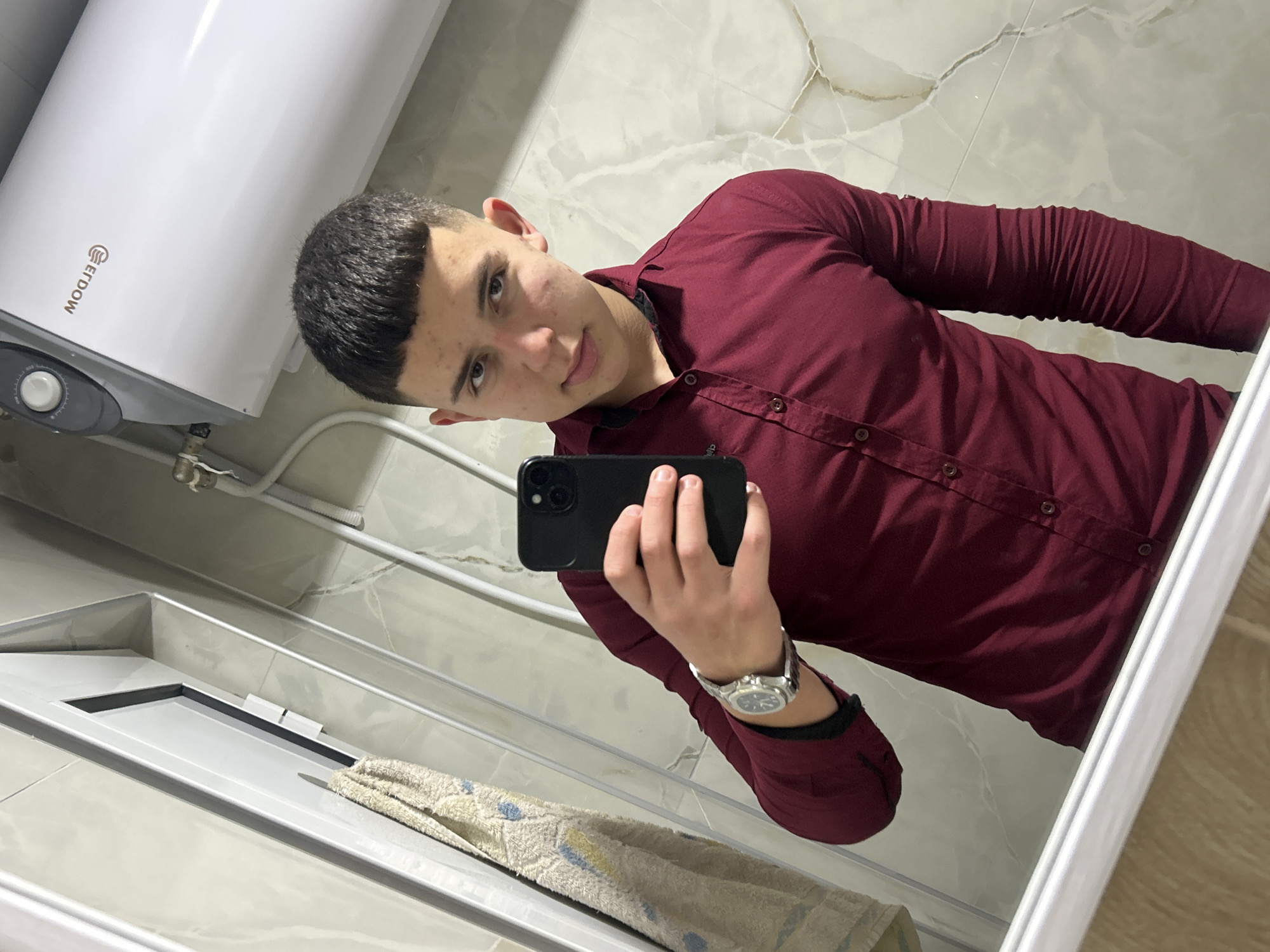 A person in a burgundy button-down shirt holds a black smartphone in their left hand, likely taking a selfie in a bathroom or lavatory. The background shows a white water heater labeled "ERDOW" on the left side of the frame and a marbled wall on the right. A towel hangs near the bottom left corner, below a sink. The image appears to be taken inside a mirror. There is no information to suggest that the location is the city of Zlatograd, Bulgaria, or anywhere else.