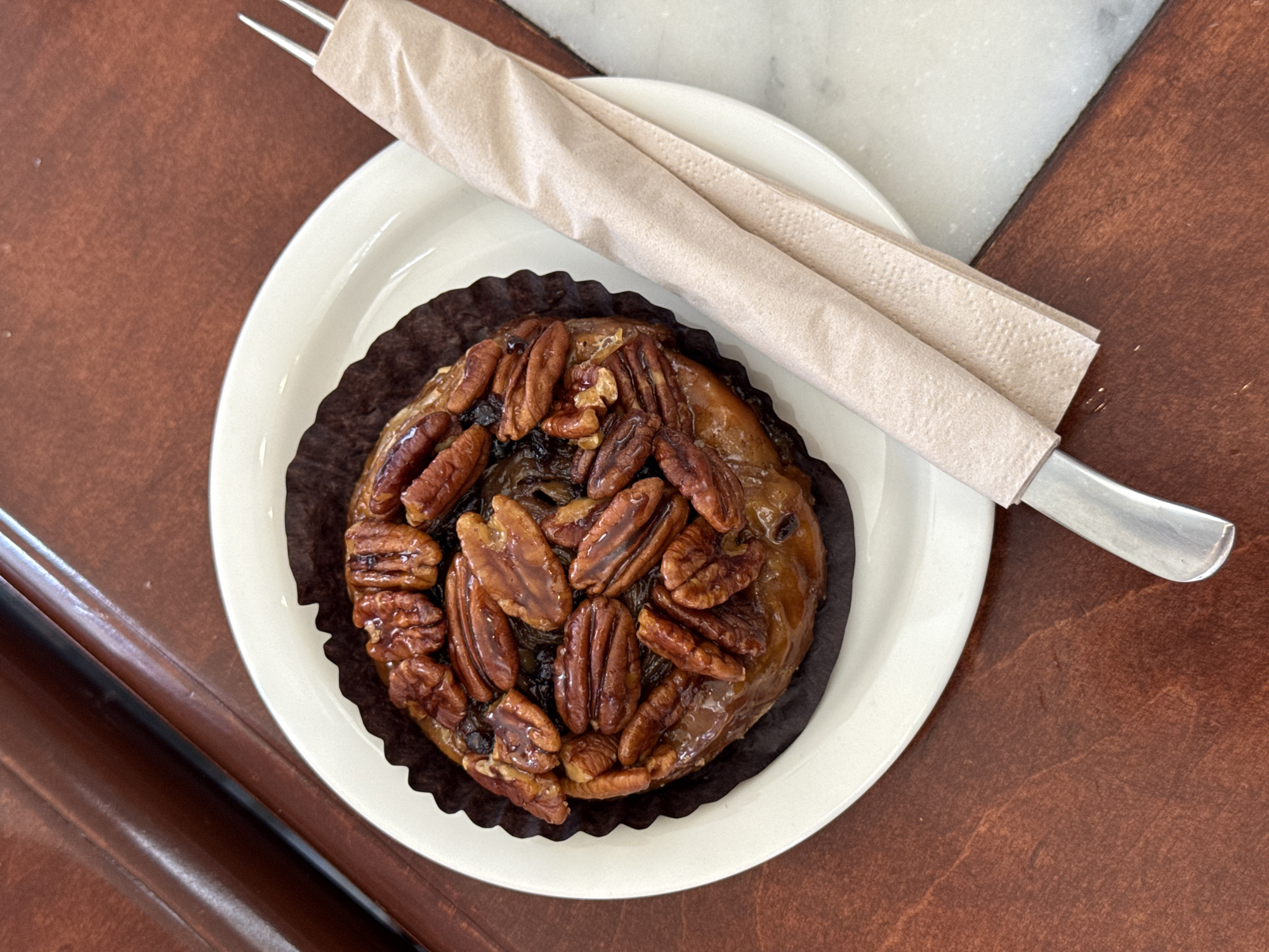 An overhead shot depicts a pecan pie on a white plate with a dark brown crinkle paper liner, a folded paper napkin with silverware resting on top, on a brown wooden table in what appears to be Beverly Hills, California. The pie is topped with pecans that are shiny and appear to be coated in a glaze. The napkin is a light tan color, folded lengthwise, and has silverware attached or nestled inside. The table is a medium brown with wood grain visible. The overall lighting is bright and natural.