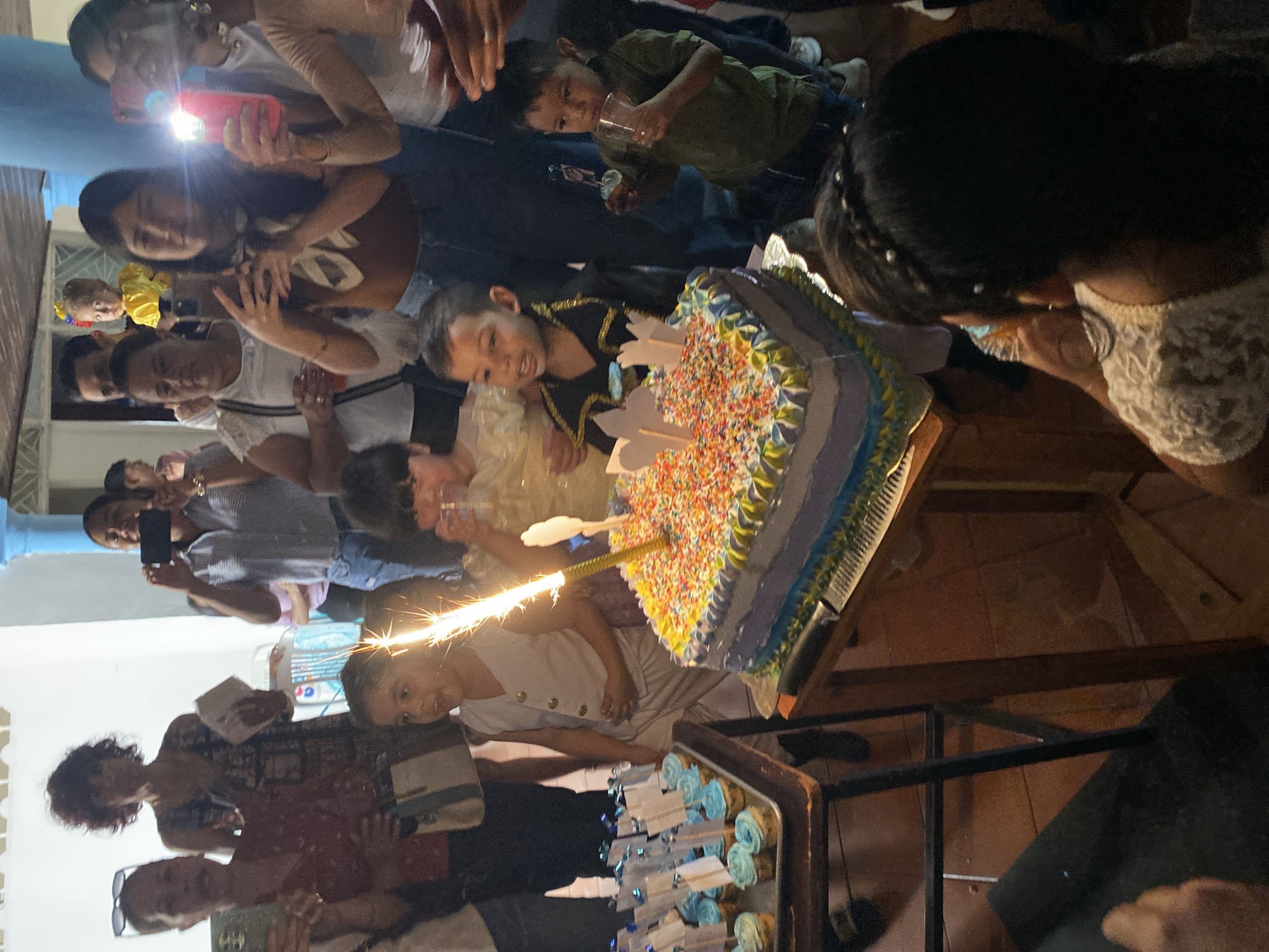 The scene depicts an indoor birthday celebration taking place in Tinaquillo, Venezuela. The central focus is a multi-layered cake positioned on a light wooden table. The cake features blue and white frosting, a top surface densely covered in multi-colored sprinkles, and is adorned with several small white, flag-like cardboard decorations. A tall, slender sparkler candle is actively emitting bright sparks from the center of the cake.
To the left of the main cake, on a separate, lower dark wooden table, sits a metal tray holding multiple cupcakes. These cupcakes are topped with blue frosting and each features a small white flag decoration.
Approximately thirteen individuals, comprising both adults and children, are visible around the cake. In the foreground, a young boy in a light-colored vest and pants stands to the left of the cake, his mouth open, looking directly at the sparkler. To the right of the cake, another young boy in a black top with gold trim observes the cake. A third young boy in a green shirt, positioned on the far right, holds a clear plastic cup and a cupcake, appearing to consume its contents. Partially visible in the bottom right, the back of a person with dark hair styled with braids and small white beads, wearing a white lace top, is directed towards the cake.
In the mid-ground and background, multiple adults are present. Several are holding cell phones, many with active camera flashes, indicating they are recording or photographing the event. A woman with dark, curly hair in a patterned dress is visible in the background. Another woman holds a phone and a light-colored shoulder bag. A small child in a yellow outfit, featuring a ribbon with the Venezuelan flag colors, is held by an adult further in the background.
The environment includes a light-colored wall in the background, contrasting with a prominent blue cylindrical pillar. The floor is tiled with light brown square tiles. Various clear plastic cups are held by attendees.