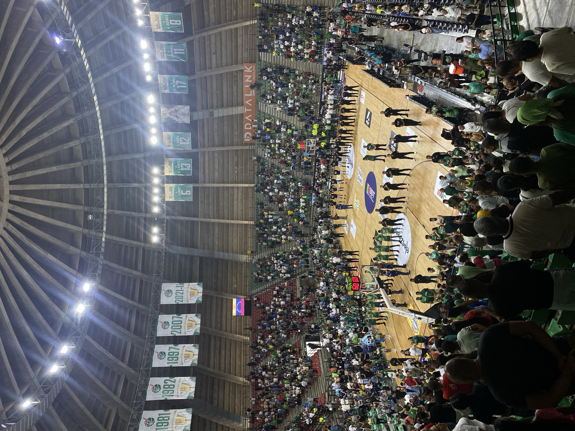 This is a photograph taken inside a crowded basketball arena in La Asuncion, Venezuela. The image is oriented vertically. The court is visible, surrounded by spectators mostly wearing green or white clothing. Banners hang from the ceiling displaying years and names, possibly indicating past championships or honored players. A scoreboard displays a time of "03:24" and a score of "60" for one team. Players, dressed in uniform, stand in rows on the court, possibly awaiting a game or participating in a ceremony. The arena's architecture includes a domed ceiling with radial supports. A small Venezuelan flag is visible among the spectators.