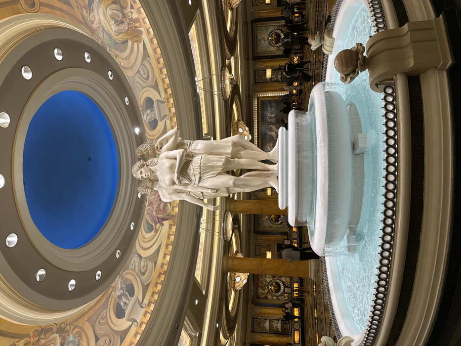 A majestic fountain with a classical sculpture stands inside a grand building. The sculpture depicts three figures atop the fountain, with cascading water flowing from tiered bowls. Above, a rotunda features a blue center surrounded by concentric rings adorned with artwork and lights. Ornate columns and chandeliers add to the building's opulent interior.