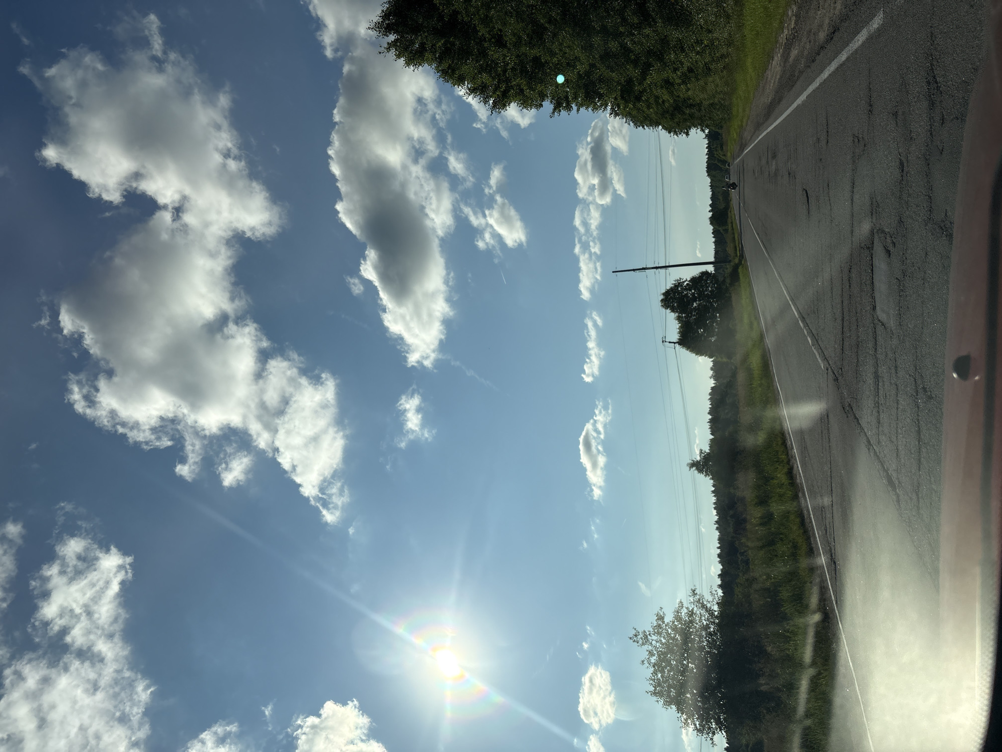 A bright sun shines through a blue sky dotted with white clouds, casting a rainbow glare on the left side of the image. The sun's rays streak across the sky. Below the sky, a two-lane asphalt road stretches into the distance, its surface cracked and uneven with numerous potholes. A white line marks the left edge of the road, and a broken white line divides the two lanes.
On the left side of the road, tall green grass and scattered trees line the shoulder. Further back, a dense forest stretches across the horizon. On the right side of the road, a line of tall green trees creates a natural border. In the distance, a motorcycle with two riders is visible on the road, heading away from the camera. Two utility poles stand along the right side of the road in the mid-ground. The foreground shows the dashboard of a vehicle, implying the photo was taken from a car. The environment appears to be rural, consistent with the location in Zamost'e, Russia.
