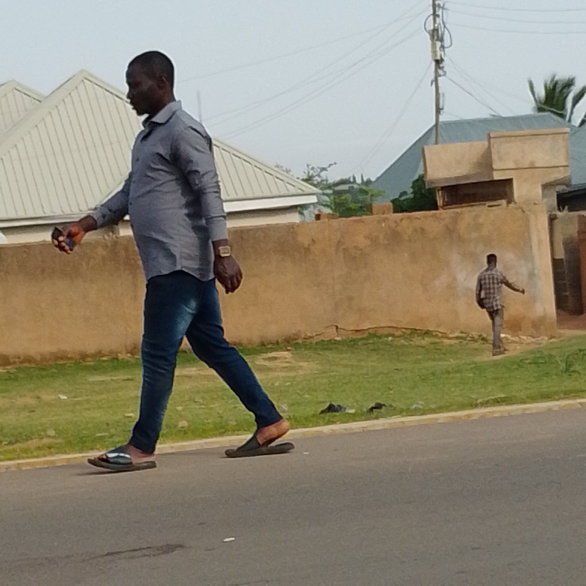 In Gombe, Nigeria, a full-body shot captures a man walking on a paved road. He is wearing a blue-grey collared shirt, dark blue jeans, and dark sandals. The man is holding a dark-colored object, possibly a phone, in his left hand. A second, smaller man in a plaid shirt and grey pants is visible further down the road, facing away from the camera and with his right arm extended. Behind the figures, a tan-colored wall runs horizontally, with a grassy area in front of it. Buildings with angled roofs and a utility pole with wires are present in the background. The sky is overcast.