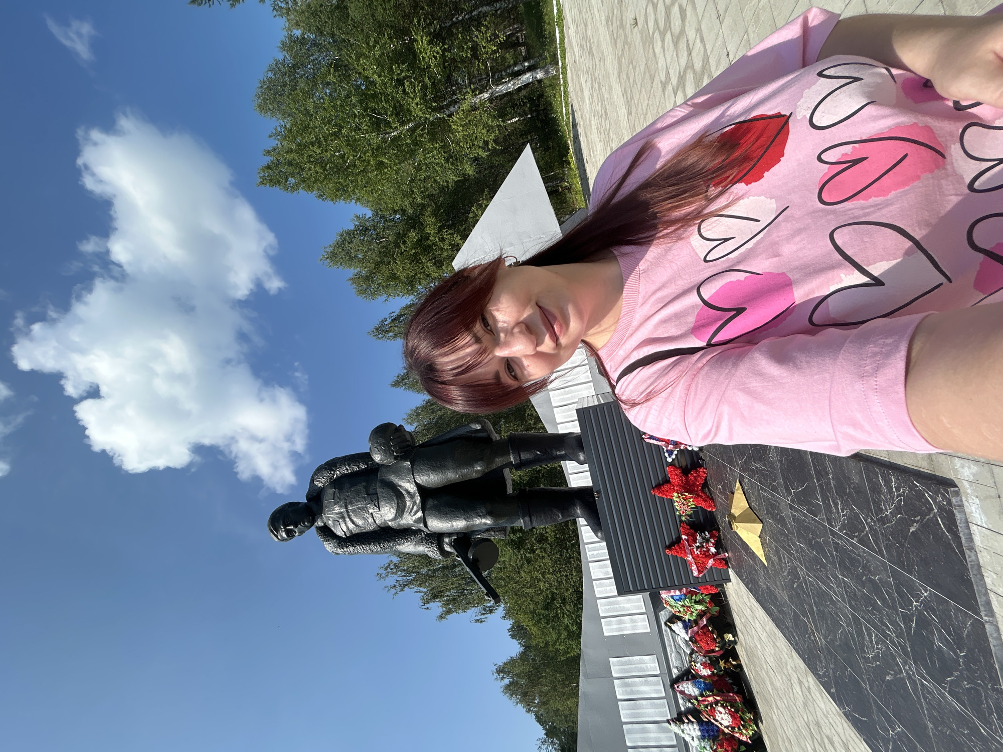 A woman with long, dark red hair, wearing a pink t-shirt adorned with heart patterns, takes a selfie in front of a memorial. The memorial features a bronze statue of a soldier holding a rifle and a child, standing on a black pedestal. Behind the statue, a gray wall with rows of inscribed names is visible. Several wreaths and red star-shaped decorations are placed at the base of the memorial. The scene is set outdoors with a clear blue sky and lush green trees in the background. The ground is paved with gray and beige square tiles. This memorial is located in the city of Krasnovishersk, Russia.