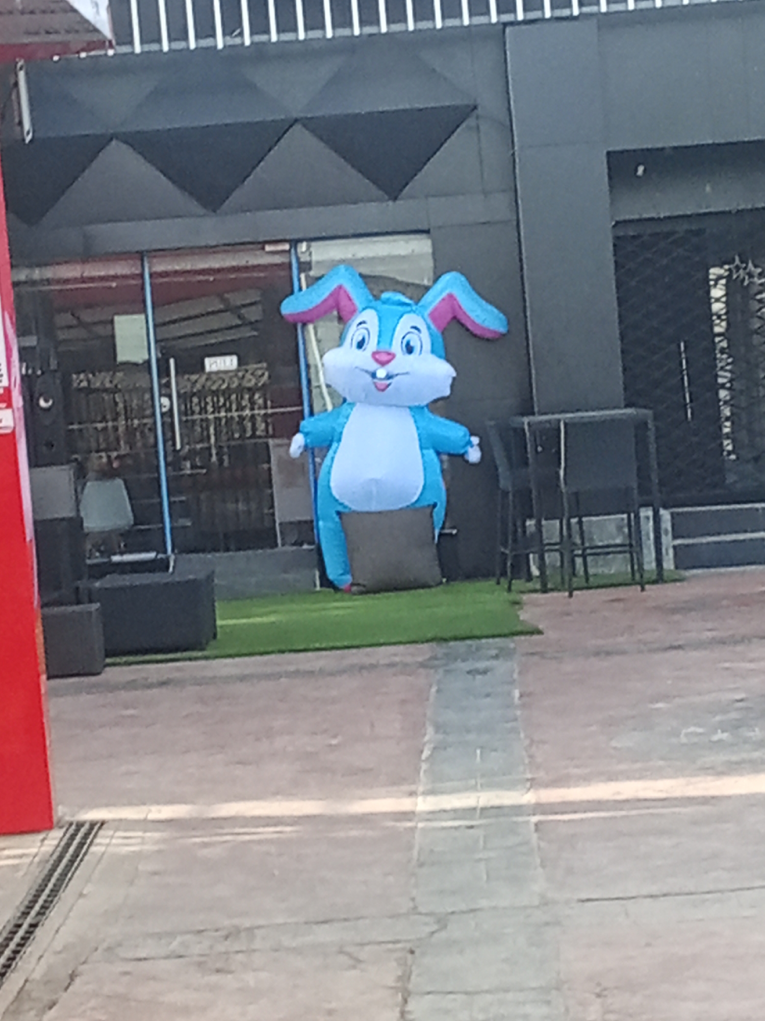 An inflatable cartoon bunny stands on a patch of artificial turf in front of a commercial building. The bunny is blue and white, with pink accents on its ears. Behind the bunny are two doors, one glass and one metal with a decorative metal design. A small seating area is visible to the left of the bunny. The building has a dark grey exterior with a striking angled design above the glass door. The ground in front of the bunny and building is paved with grey bricks. This image could be from Ado, Nigeria.