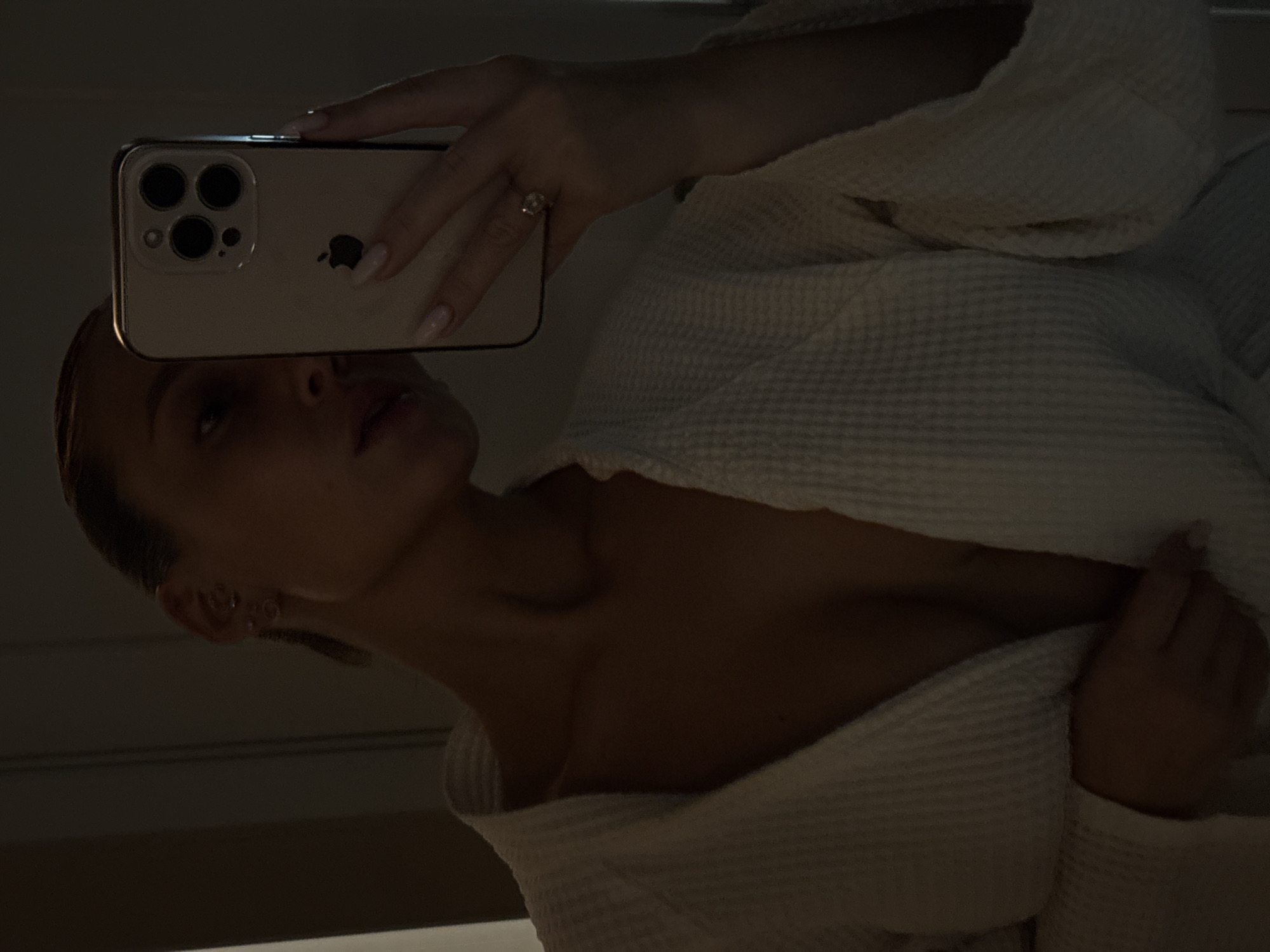 A woman in a white robe takes a selfie with a phone. The phone is held in her right hand, and it covers the upper half of her face. She wears earrings, and a ring is visible on her right ring finger. The background is dark and indistinct. There is no information to determine if the location of the image is Chaikovsky, Russia.