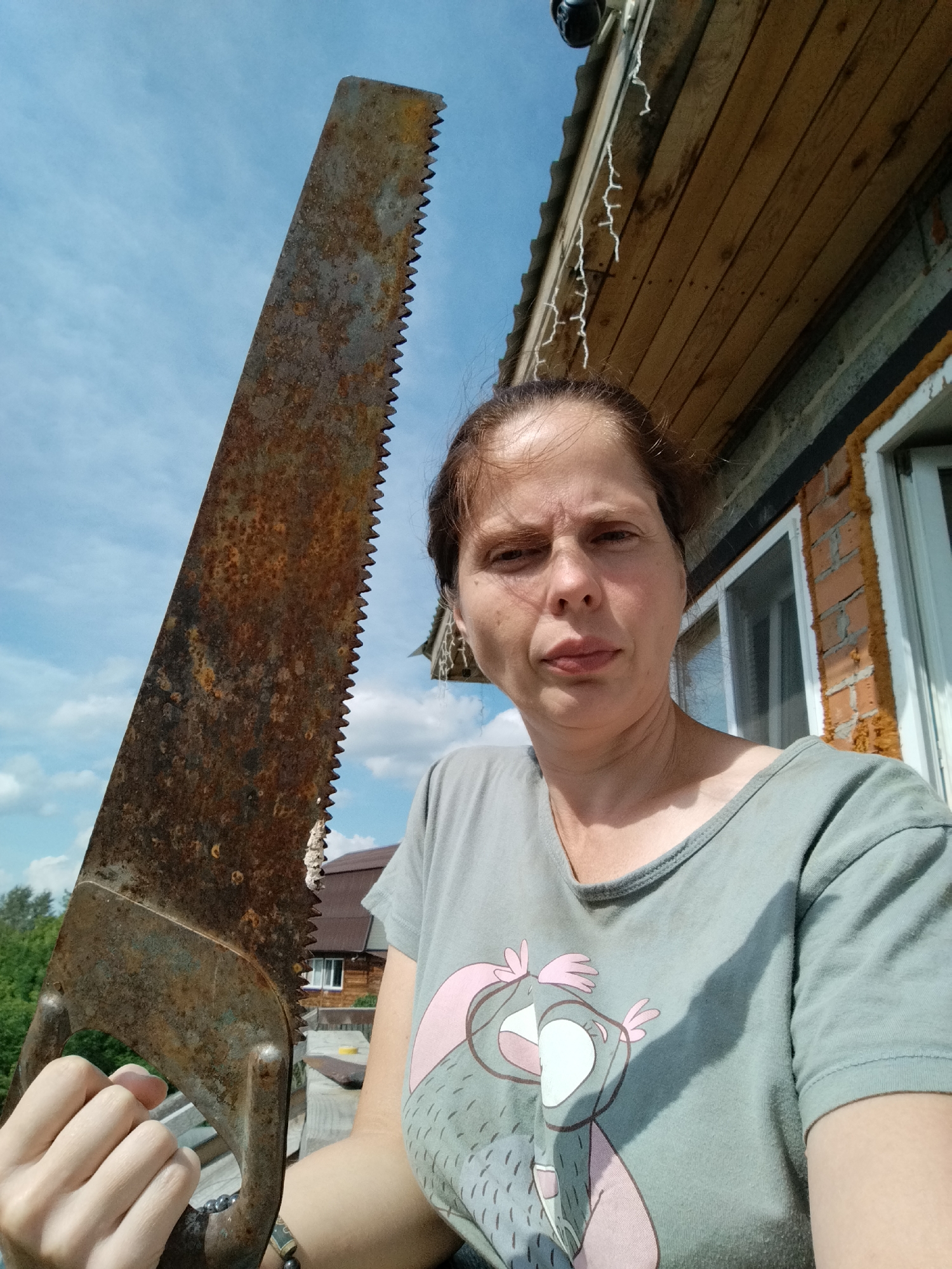 A person is holding a rusty saw in front of a building with a wooden roof and brick walls. The person is wearing a grey t-shirt with a cartoon owl print. The background shows a blue sky with clouds and some trees and houses. The scene is likely outdoors.