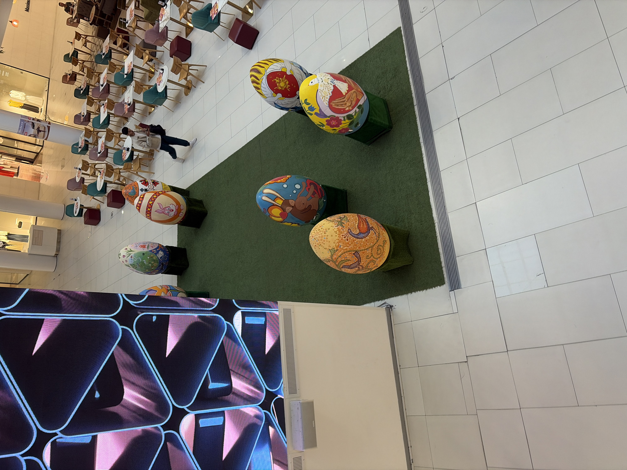 The image shows a mall setting, likely in Plovdiv, Bulgaria, based on common mall design and decoration practices. Oversized, decorated Easter egg sculptures sit on small grass patches. A woman walks among tables and chairs, potentially part of a mall restaurant. A large digital display showing a pattern of triangles is visible in the foreground. The flooring consists of white tiles, and the overall lighting suggests an indoor, artificial source.