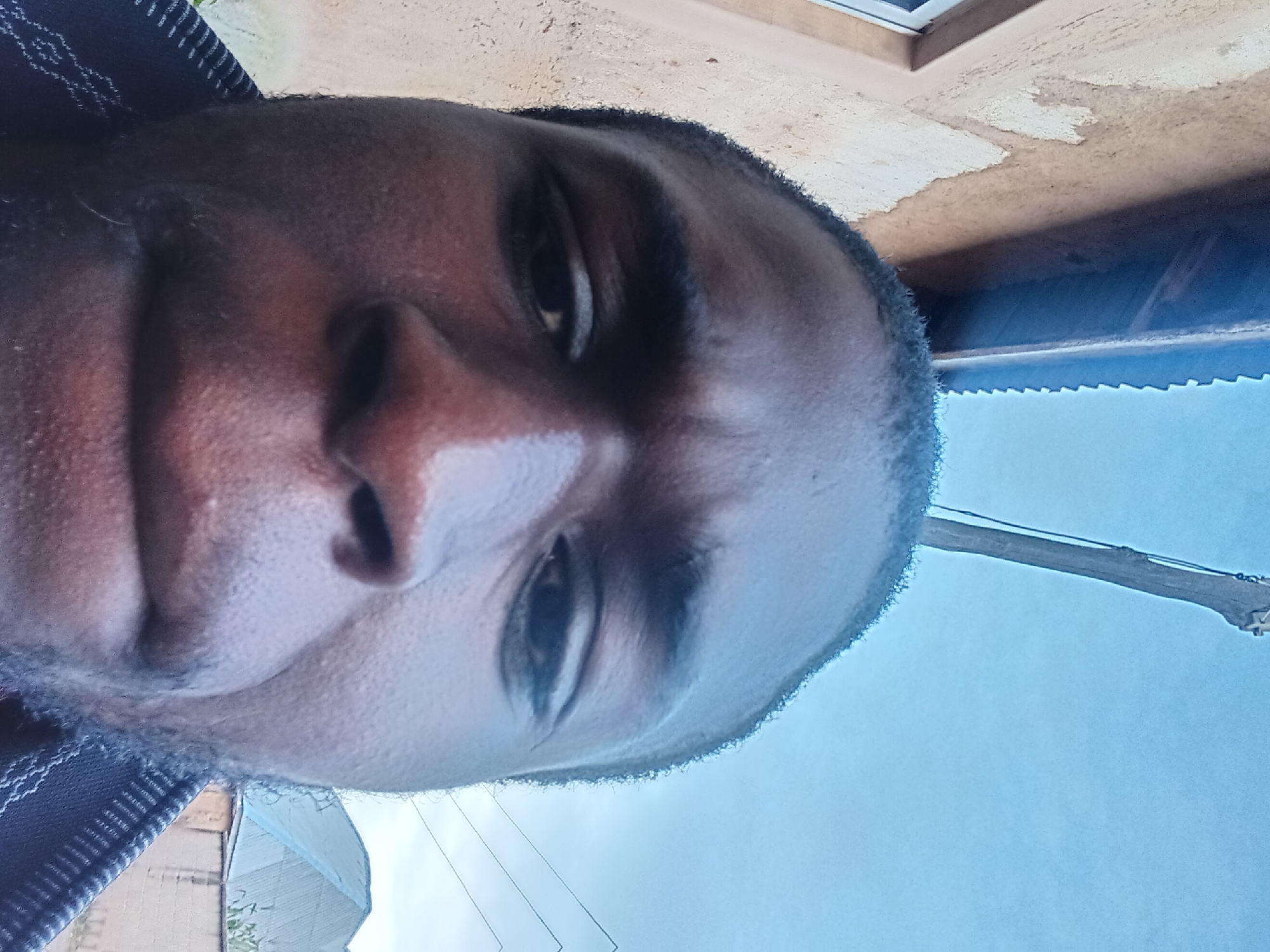 A close-up, frontal, eye-level, low-angle selfie taken outdoors in Bukuru, Nigeria. The image features a dark-skinned man from the chest up, his face filling most of the frame. He has dark hair and is wearing a dark patterned garment around his neck and shoulders. The man's expression is neutral, with his mouth slightly parted. In the background, a beige building with a blue corrugated metal roof is visible to the left, and a light blue sky dominates the upper portion of the image. A thin, upright wooden pole is also present in the sky. A residential building with a blue roof and a brick wall can be seen in the background to the right.