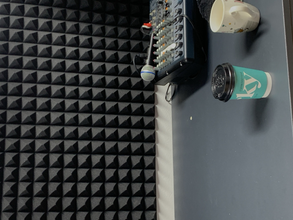 The image depicts an indoor studio environment. The background features a wall entirely covered with black, pyramid-shaped acoustic foam panels, with a narrow white wall section visible at its base. A dark grey desk surface occupies the foreground. On the right side of the desk, an audio mixing console with multiple knobs and faders is positioned, accompanied by a microphone with a blue and white windscreen resting nearby. Several electrical cables are connected to the mixer. In the foreground, a turquoise paper coffee cup with a black lid displays the text "EXPRESSO BAR" and "SKY" with a prominent "KY" visible in its branding. To the right of the paper cup, a white ceramic mug adorned with colorful designs is present. No individuals are visible. The "KY" branding on the paper cup indicates the location is in Kyiv, Ukraine, likely within a studio or facility that sources beverages from a "Sky Espresso Bar" establishment in the city.