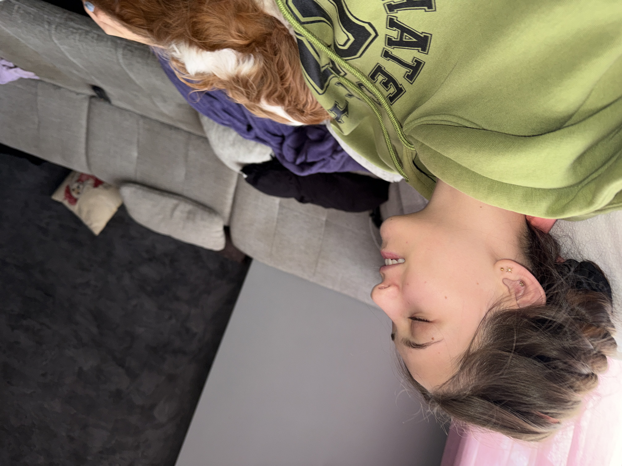 A woman lies on a gray sofa. She wears a green sweatshirt with black lettering. A brown and white dog sits beside her. The location is not specified in the image or recognizable from any visual cues as being in Sofia, Bulgaria. The sofa has several pillows. The background is a gray wall.