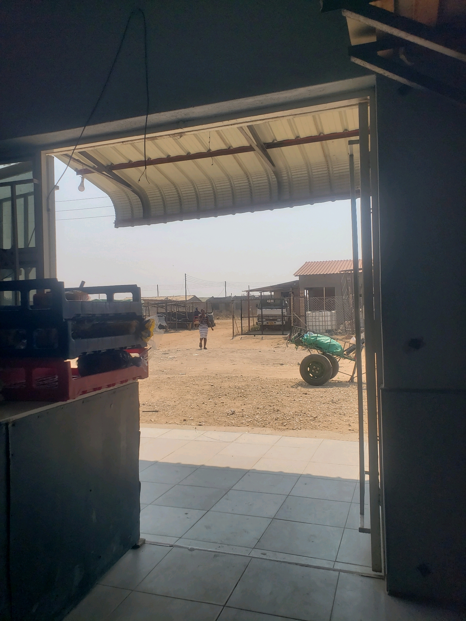 The image depicts an outdoor scene, likely in a dusty, rural or semi-urban area, possibly in or near Masvingo, Zimbabwe. The foreground shows the interior of a structure, with tiled flooring and a counter or dark-colored surface on the left. A doorway or opening leads to the outside.
Outside, a person is walking away from the camera, wearing a striped shirt and shorts. The ground is dirt and gravel. In the mid-ground, there are several structures with corrugated metal roofs, some appearing to be stalls or small buildings. A white truck is parked near one of these structures. To the right, a green-covered cart with two large wheels is visible, loaded with some items. The environment is bright and appears to be under a clear sky, suggesting daylight. The overall impression is of a marketplace or an area with small businesses.