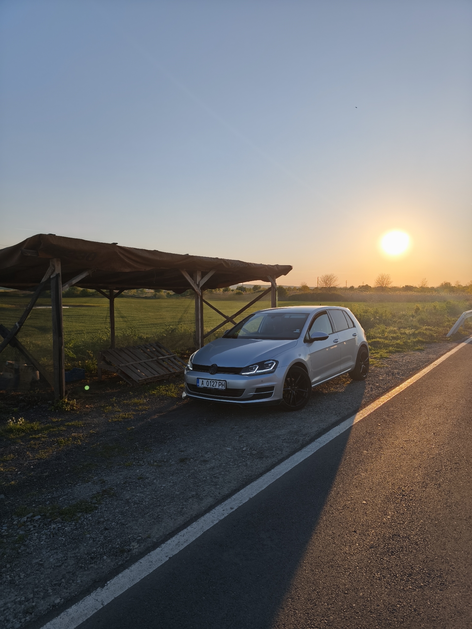 The image shows a silver Volkswagen Golf parked near a small, wooden shelter with a tarp roof next to a road. The car has a Bulgarian license plate "A 0127 PH." The sun is setting in the background, casting a warm glow over the scene. The location appears to be rural, with a field visible behind the shelter. The road has a white dividing line. This is in the Trastikovo region of Bulgaria.