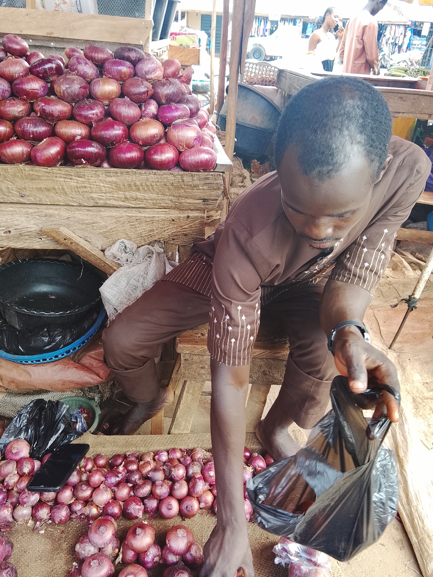 A market scene in Akwanga, Nigeria, features a man in a brown outfit with white vertical stripes on the sleeves, reaching down towards a pile of red onions. He is holding a black plastic bag, appearing to be preparing to put onions into it. A smartphone is visible on top of a pile of onions to the left. In the background, other individuals are present, along with market stalls displaying various goods, including clothing and produce. The environment suggests a typical outdoor market setting with wooden structures and a packed ground.
