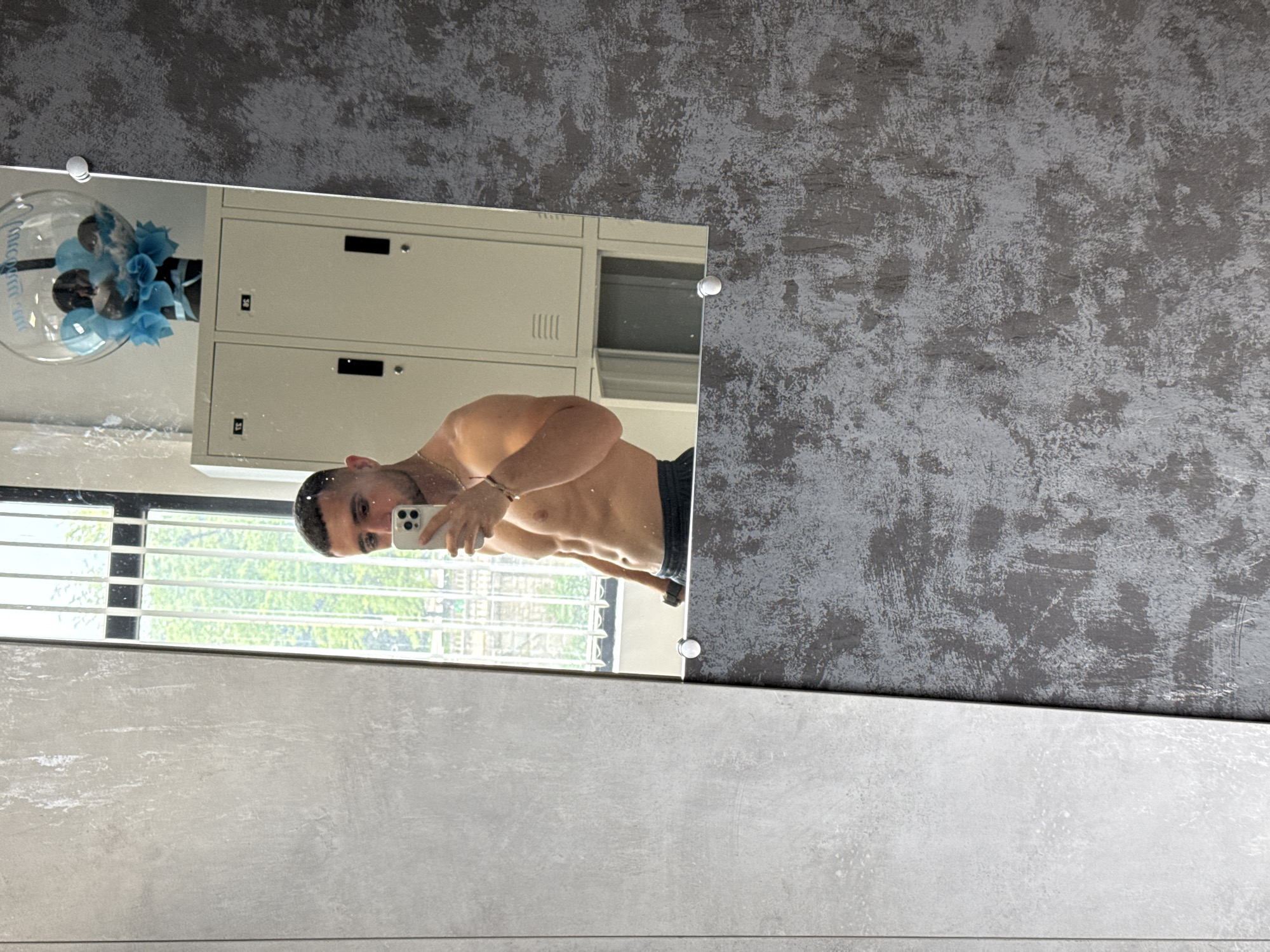 A shirtless man is taking a selfie in front of a mirror. The mirror is attached to a wall with a grey, textured upper portion and a lighter grey, smooth lower portion. The reflection in the mirror shows a window with white blinds, a white locker, and a decorative floral arrangement in a clear bowl. The location appears to be indoors, possibly a locker room or bathroom. Gabrovo, Bulgaria is not discernible from the image.
