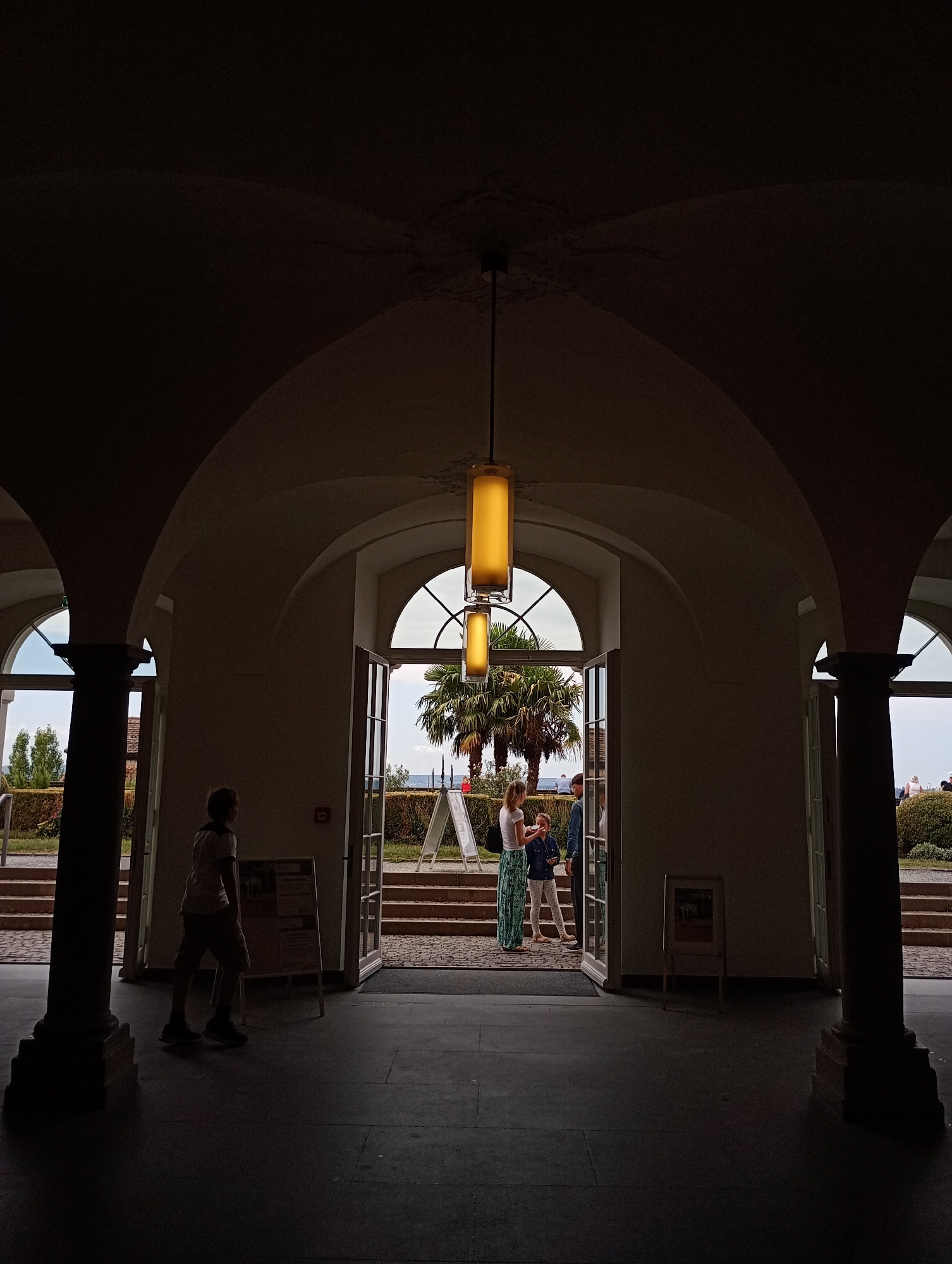 The image depicts an interior view of a building with arched ceilings and columns, looking out towards an exterior landscape. In the foreground, a young person, silhouetted against the light, walks away from the viewer towards the left. They are wearing a t-shirt and shorts. Further back, a woman in a long, patterned skirt and a light-colored top, and a child wearing a blue jacket and white pants, are standing near an open doorway. They appear to be interacting. Beyond them, a palm tree is visible, with stairs leading up to a garden area with more trees and shrubs. The exterior also shows a body of water, likely Lake Constance, and distant hills. There are several A-frame signs visible both inside and outside the building. The lighting suggests it is daytime, with strong backlighting from the exterior creating silhouettes of the interior architecture and people. This scene is located at the entrance to the Meersburg Castle (Alte Schloss Meersburg) in Meersburg, Germany.