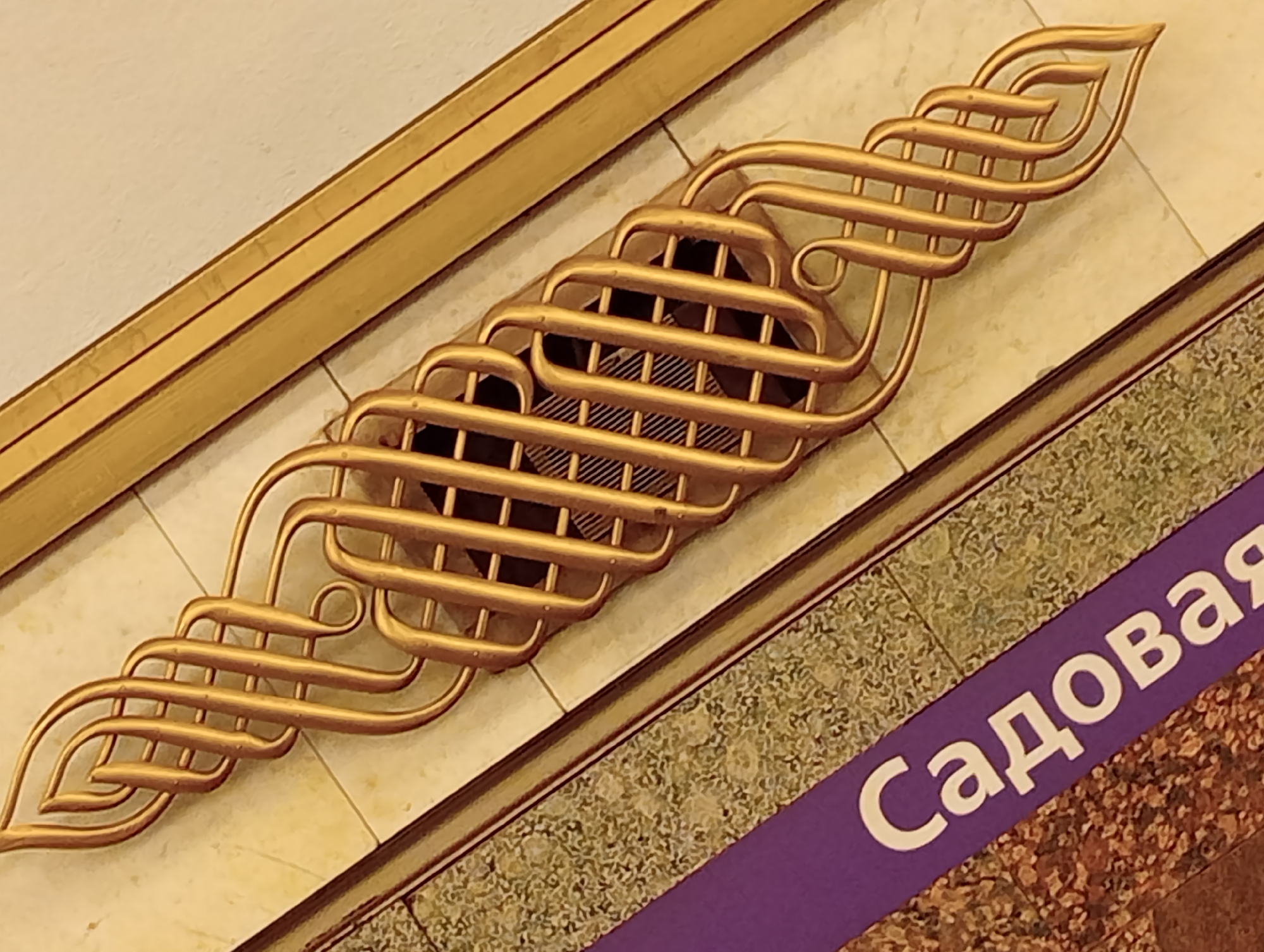 The image shows a detail of the decorative design and signage at a metro station in Saint Petersburg, Russia. The main focus is a golden, stylized wing-like ornament made of curved metal tubes, superimposed over a dark grille. This ornament is positioned diagonally across the frame, with its left and right ends extending outwards. The background consists of light-colored tiles. Below the ornament, a purple band runs diagonally, bearing white Cyrillic lettering that reads "Садовая" (Sadovaya). This signage indicates the name of the station. The lower portion of the image reveals a textured surface, possibly granite, with flecks of different colors. The overall impression is one of ornate architectural detailing common in the Moscow or Saint Petersburg Metro systems, likely from the Soviet era or a subsequent renovation.