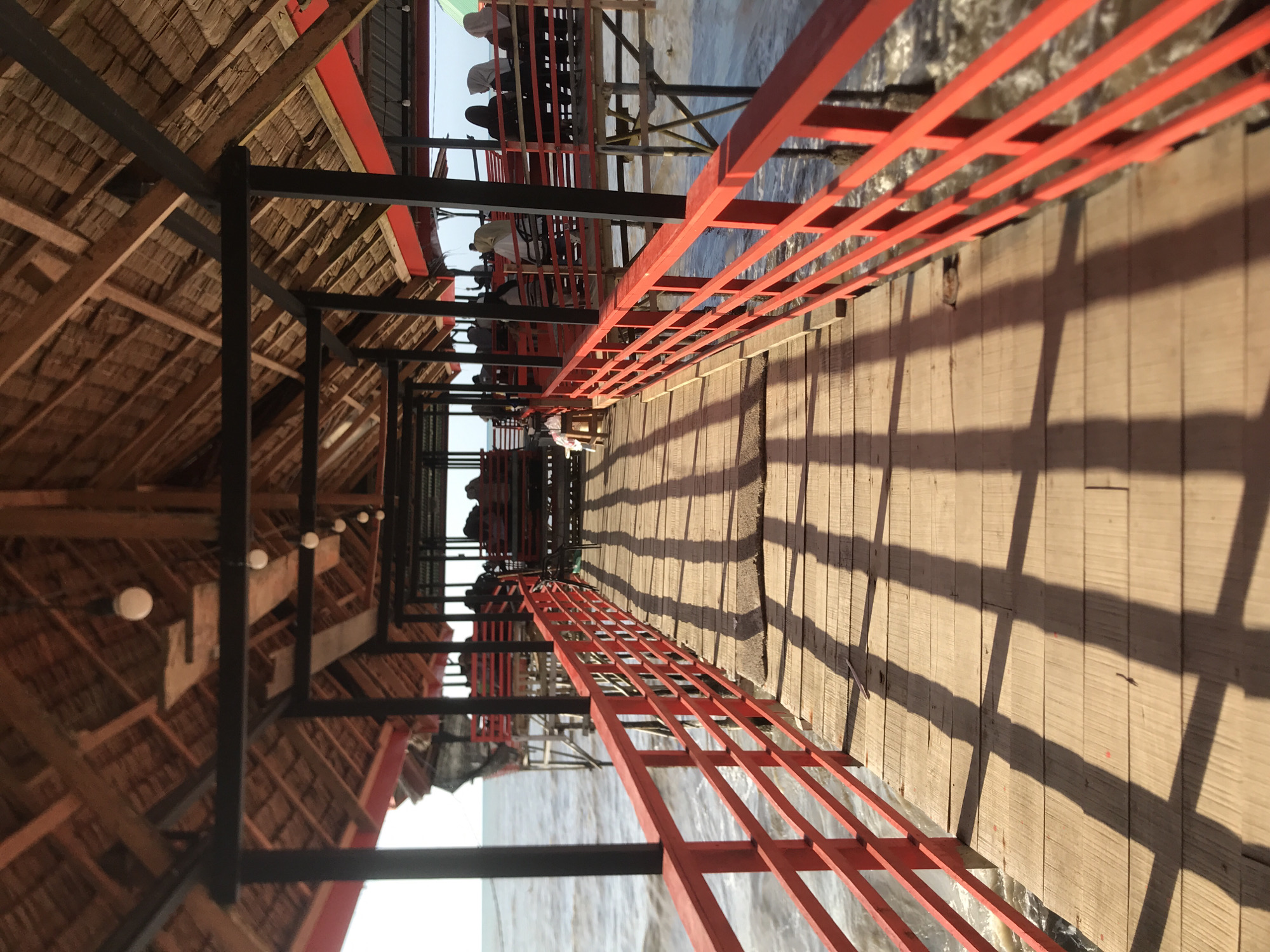 The image depicts a long, covered boardwalk extending over water, likely a lake or river, in Lancok, Indonesia. The boardwalk has a thatched roof and is supported by black metal beams. Red metal railings line both sides of the boardwalk. People can be seen in the distance, seated at tables under an open-air structure at the end of the boardwalk. The boardwalk itself is made of light-colored wood, with distinct shadows cast by the railings. The water is visible beneath the boardwalk.