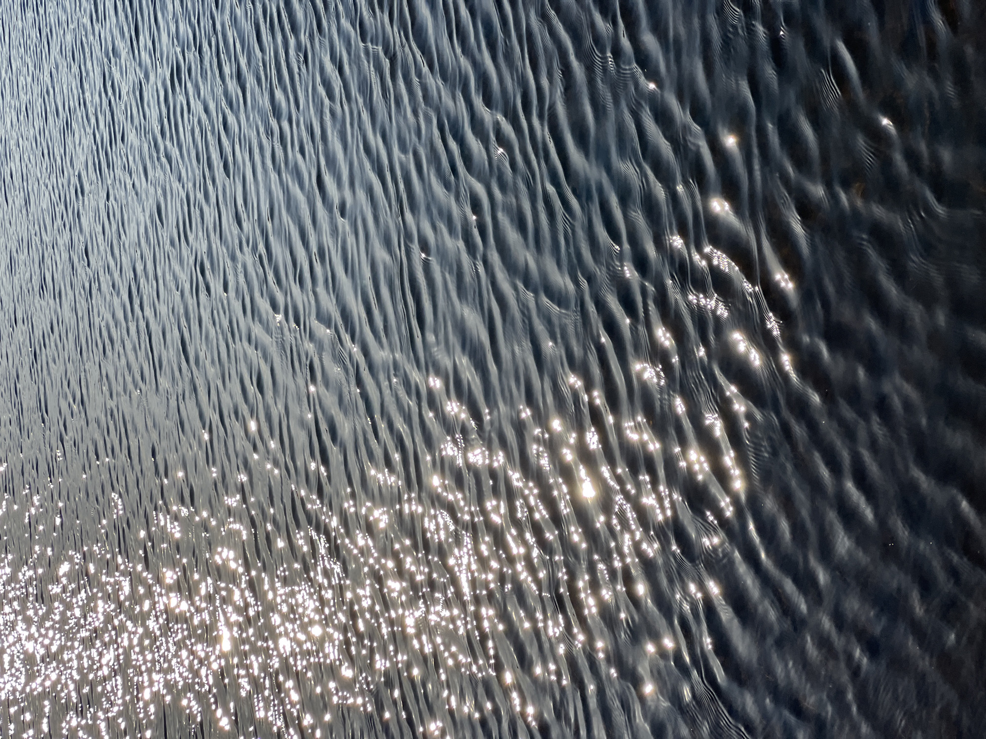 The image shows the surface of the water in the city of Chaikovsky, Russia. The water has small, regular ripples, and the sun reflects brightly off the surface in a scattered pattern. The overall color of the water is a dark grey-blue, with the reflected light appearing white.