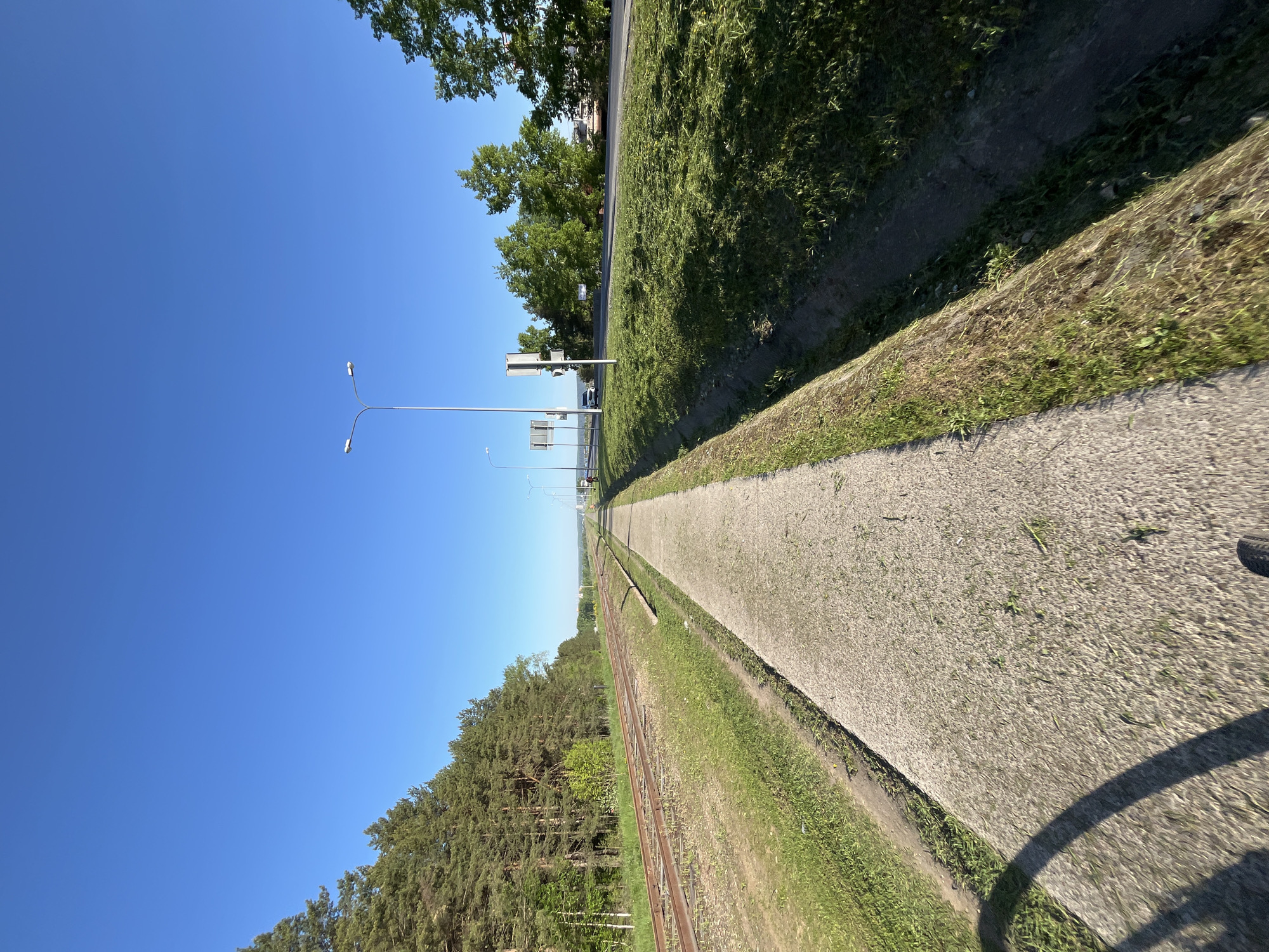 The image shows a view along a section of train tracks in Chaikovsky, Russia. The tracks run toward the horizon between a gravel path on the right and a grassy area leading to a forest on the left. On the other side of the tracks are tall grass, trees, and a road with streetlights and traffic signs in the distance. The sky is clear and blue. A shadow of a wheel is in the bottom right corner, indicating the photograph was taken from a moving vehicle or bicycle.