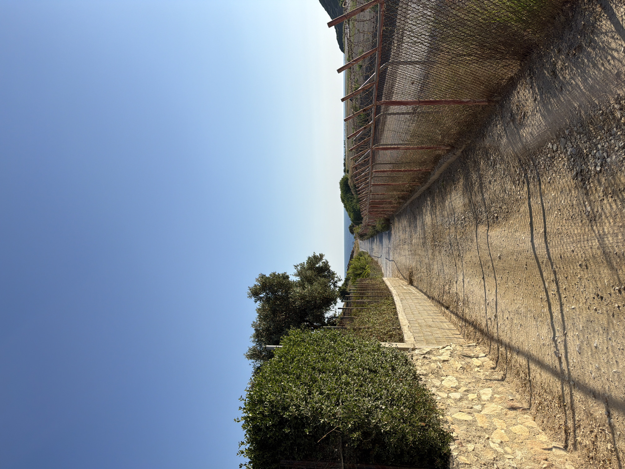 The image depicts a dirt path leading towards the sea under a clear blue sky. On the left, a stone wall topped with dense green foliage separates the path from a rocky area. Further down the path, a chain-link fence with red posts and barbed wire runs along the right side. The path itself shows tire tracks and scattered stones, indicating it is likely a service road or trail. In the distance, the sea is visible, bordered by low green hills. The overall impression is one of a coastal access road. The scene is likely located in or near Skiathos, Greece, given the typical landscape and the presence of similar fencing around airport perimeters. There are no people or animals visible in the image.
