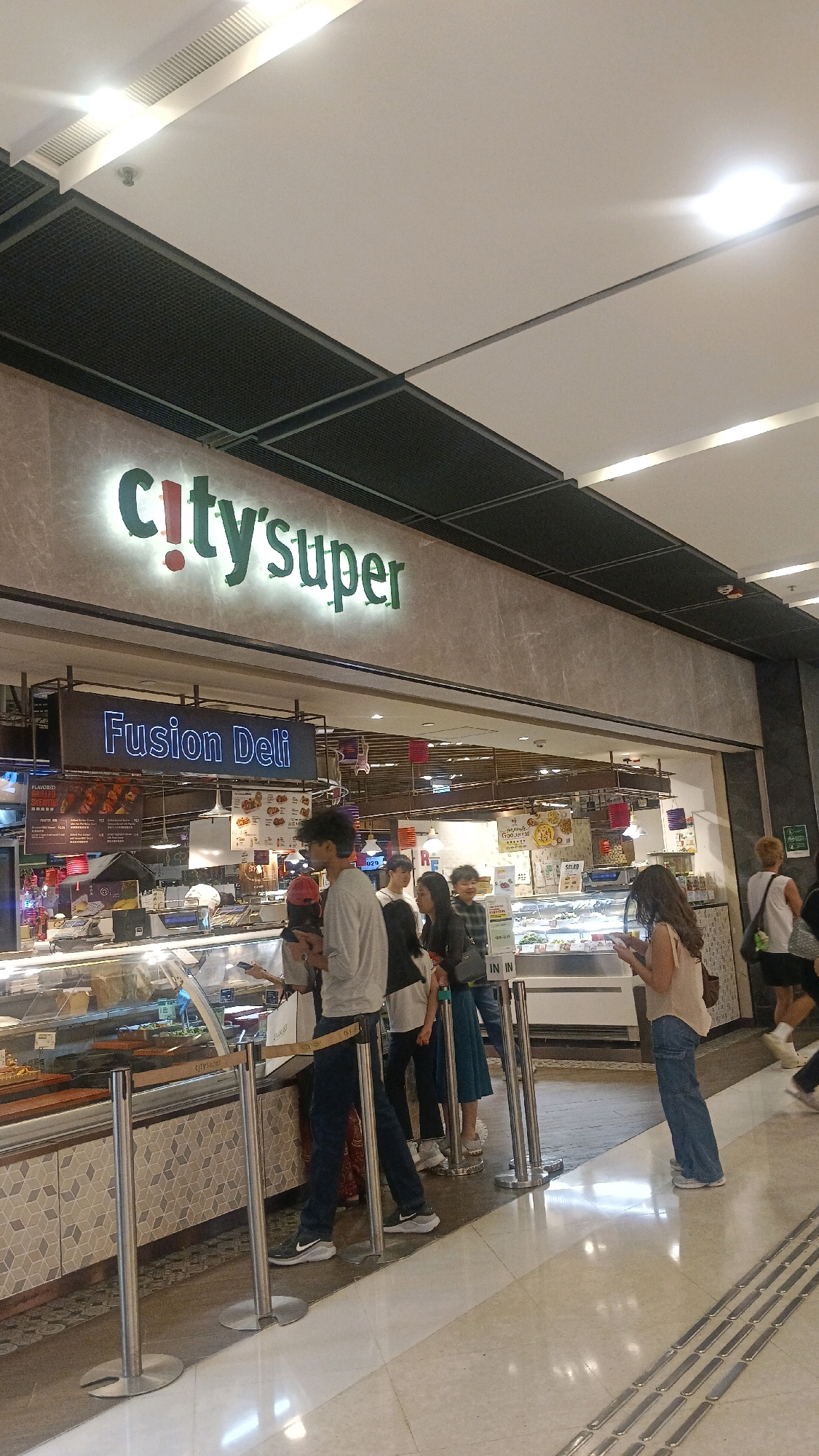 This image captures a scene inside a City'super store in Hong Kong, China. The store features a prominent "City'super" sign with illuminated green lettering. Below the main sign is a smaller sign that reads "Fusion Deli" in a blue neon font. A queue of people, consisting of young adults and teenagers, are standing in line in front of a food counter. The counter displays various food items behind glass, suggesting a prepared foods or deli section. The environment appears to be a modern, well-lit shopping mall or supermarket interior, with a tiled floor and an elevated ceiling with lighting fixtures. The people are casually dressed, and some are looking at their phones. The overall atmosphere is one of everyday commerce and consumer activity.