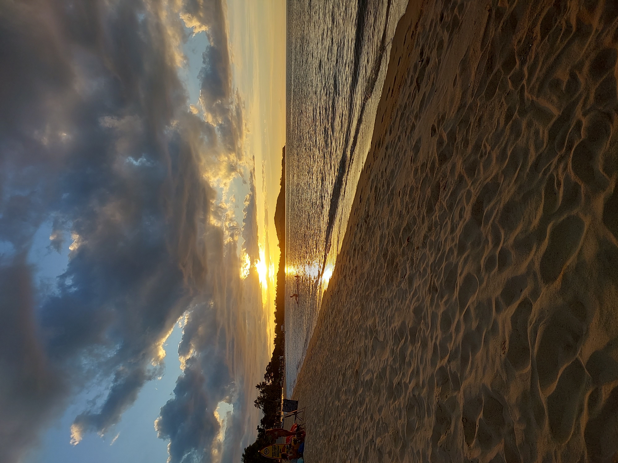 The image depicts a serene beach scene at sunset in Baan Mae Nam, Thailand. The sun, low on the horizon, casts a golden glow across the water and the sand. The sky is a mix of dramatic dark clouds tinged with orange and yellow from the setting sun, creating a beautiful contrast with the clearer patches of blue sky. The ocean is calm, with gentle waves lapping at the shore, reflecting the warm light of the sun.
The foreground is dominated by the texture of the sandy beach, with ripples and footprints creating a sense of depth and movement. In the distance, a silhouette of landmass, likely an island or mainland, can be seen against the bright horizon. A lone figure on a paddleboard is visible on the water, adding a touch of human presence to the vastness of the scene. Further down the beach, there are hints of palm trees and vegetation, characteristic of a tropical location, and a few people are gathered near some beach chairs and a surfboard, enjoying the end of the day. The overall mood is peaceful and tranquil, capturing the beauty of a tropical sunset. No visible text is discernible.