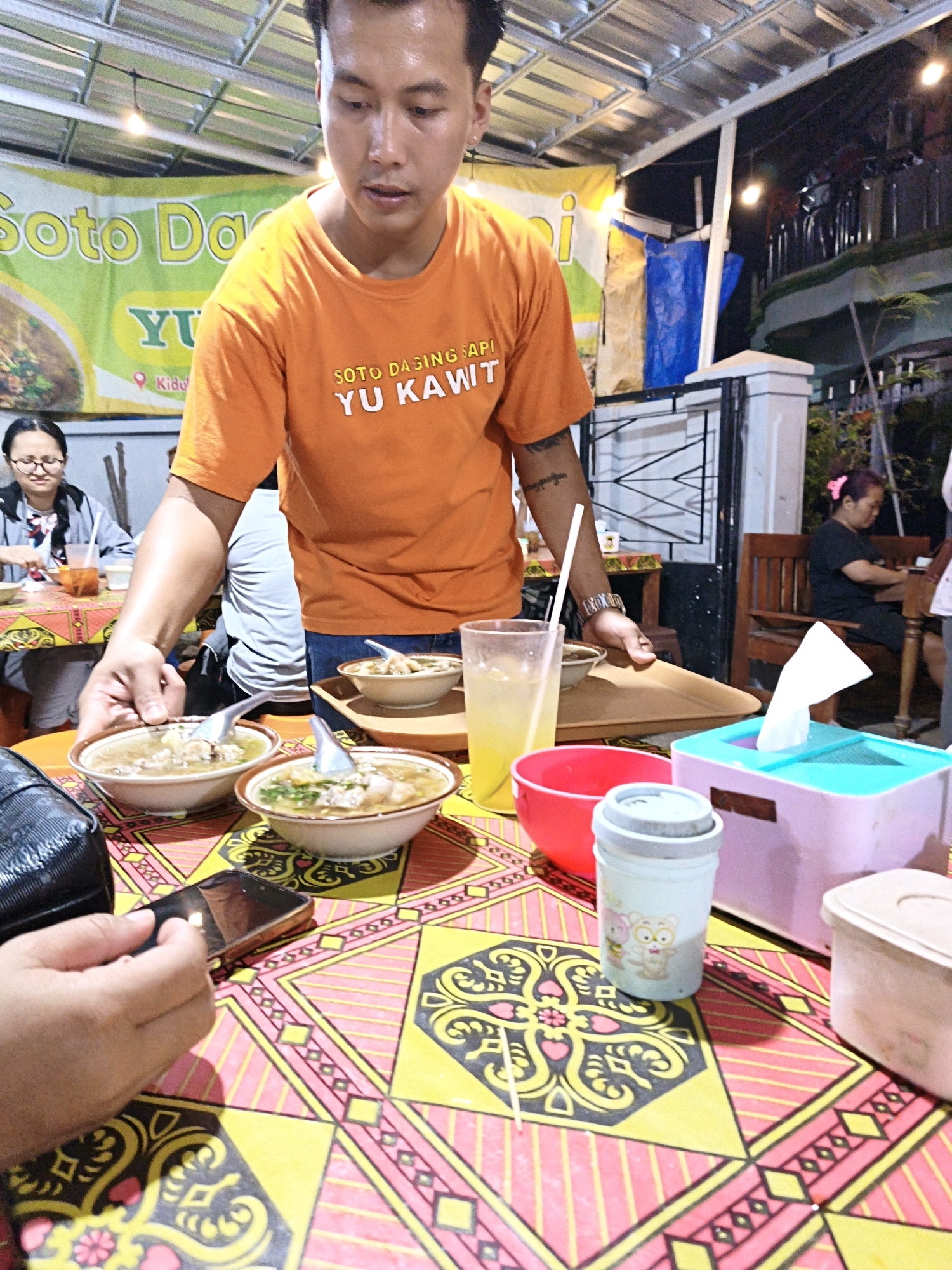 An Asian man wearing an orange t-shirt with the text "SOTO DAGING SAPI YU KAWIT" on it is serving bowls of soup. He is standing behind a table covered with a colorful cloth patterned with yellow, pink, and black geometric designs. In front of him are at least three bowls of soup, each with a spoon. To his right, a tray is visible, as well as a tall glass with a yellow drink and a straw, a red bowl, a blue and pink tissue box, and a small cylindrical container with cartoon characters. A black bag and a smartphone are on the table in the foreground, with a person's hand holding the phone. In the background, a woman with glasses is seated at a table, and another woman with purple hair is sitting on a wooden bench. The scene appears to be an outdoor food stall or restaurant, with a metal roof overhead supported by beams and illuminated by string lights. The banner behind the man indicates the food stall is "Soto Daging Sapi Yu Kawit" and shows a location marker for "Kidu". This is likely a scene in Surakarta, Indonesia, where this type of street food stall is common.