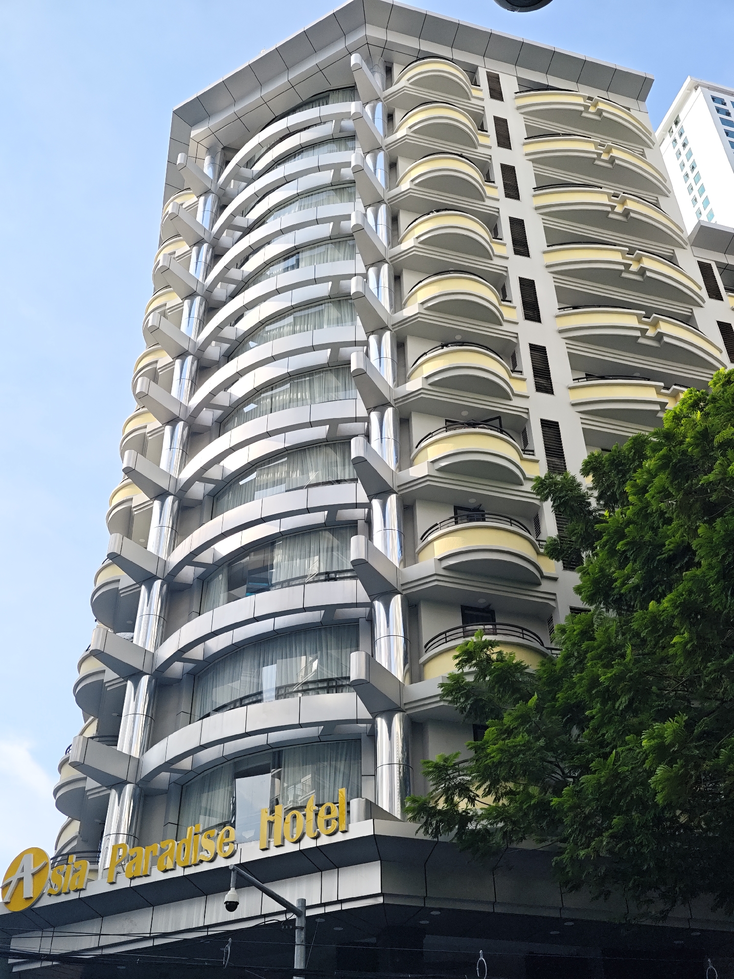 This is a low-angle, full shot of the Asia Paradise Hotel in Nha Trang Ward, Vietnam, against a clear blue sky. The hotel is a tall, multi-story building with a curved facade, characterized by a series of repeating balconies with yellow trim. The building's exterior features a mix of gray and silver paneling, with large, floor-to-ceiling windows and chrome-like cylindrical columns. The hotel's name, "Asia Paradise Hotel," is prominently displayed in large, yellow, raised lettering on the lower part of the building, accompanied by a yellow circular logo. Below the hotel's sign, a surveillance camera is visible mounted on a pole. To the right of the hotel, the lush green foliage of a large tree partially obscures the view of the building's lower floors and right side. In the background, a portion of another tall white building with visible windows can be seen. The scene depicts an urban architectural setting during daylight hours, with no people or any discernible activity.