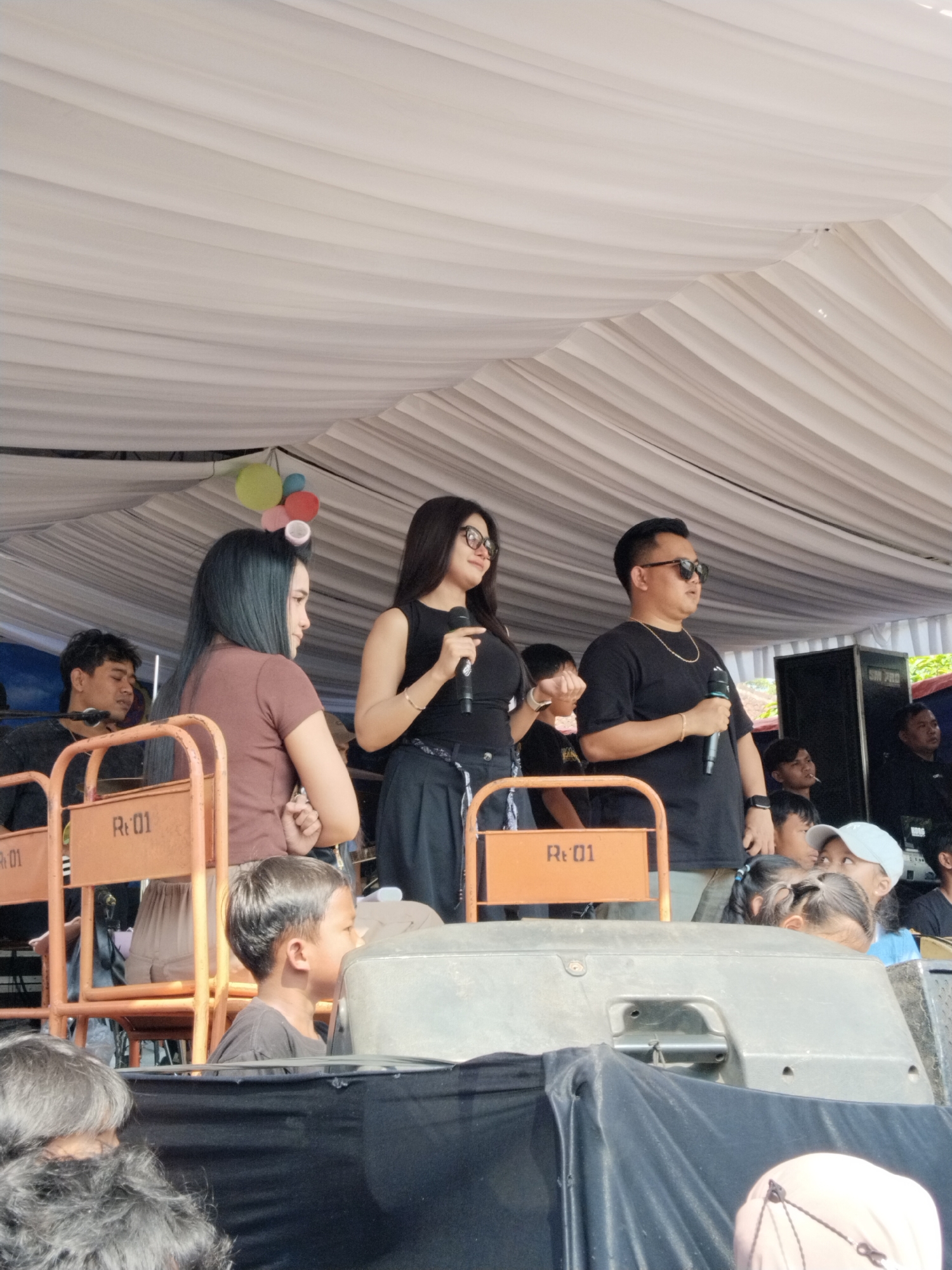 In the city of Ngadirejo, Indonesia, three individuals stand on a stage set up under a white tent. Two women and one man are positioned at the front of the stage, each holding a microphone. The woman on the left has long dark hair and wears a brown shirt. The woman in the center has long dark hair and wears a black sleeveless top and a black skirt. The man on the right has short dark hair, sunglasses, a gold chain, and wears a black t-shirt. They appear to be performing or speaking to an audience. In the foreground, a young boy in a black t-shirt is visible, looking to his right. Various objects like chairs with orange frames and the text "Rt. 01" are present. A large speaker is visible behind the man on the right, and a large white tent covers the entire area, suggesting an outdoor event. The lighting suggests it is daytime.