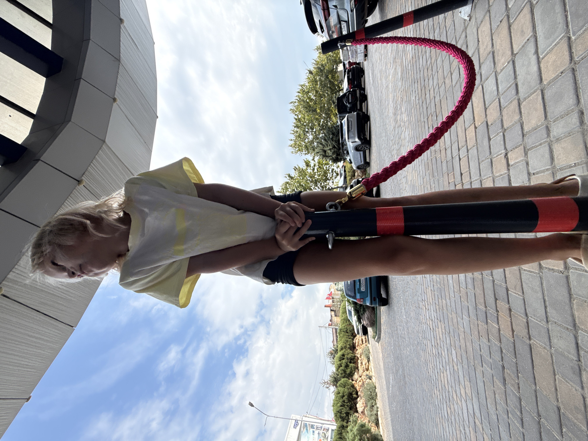 The image shows a young girl from the waist down, leaning forward. She is wearing a yellow t-shirt and dark shorts. Her hands are resting on a black pole with red accents, which is part of a stanchion with a thick red rope attached. The stanchion and rope suggest a cordoned-off area, possibly for a queue or to designate a specific zone.
The setting appears to be outdoors, on a paved walkway made of interlocking grey pavers. In the background, several cars are parked in a line, suggesting a parking lot or a street. Beyond the cars, there is a line of green trees and bushes, and further back, some buildings and a lamppost are visible. The sky is a bright blue with some white clouds, indicating it is daytime, likely late morning or early afternoon, and the weather is clear.
The girl's posture and facial expression, partially visible in the top left of the image, seem relaxed or contemplative. The overall scene is casual and appears to be a public space.
There is some text visible on a blue car in the background. On the license plate, the characters "B 320 KA" are discernible, along with some other symbols. Another sign is visible in the far background with text, but it is too blurry to read.
The perspective of the photo is from a low angle, looking upwards, which emphasizes the height of the stanchion and the sky.