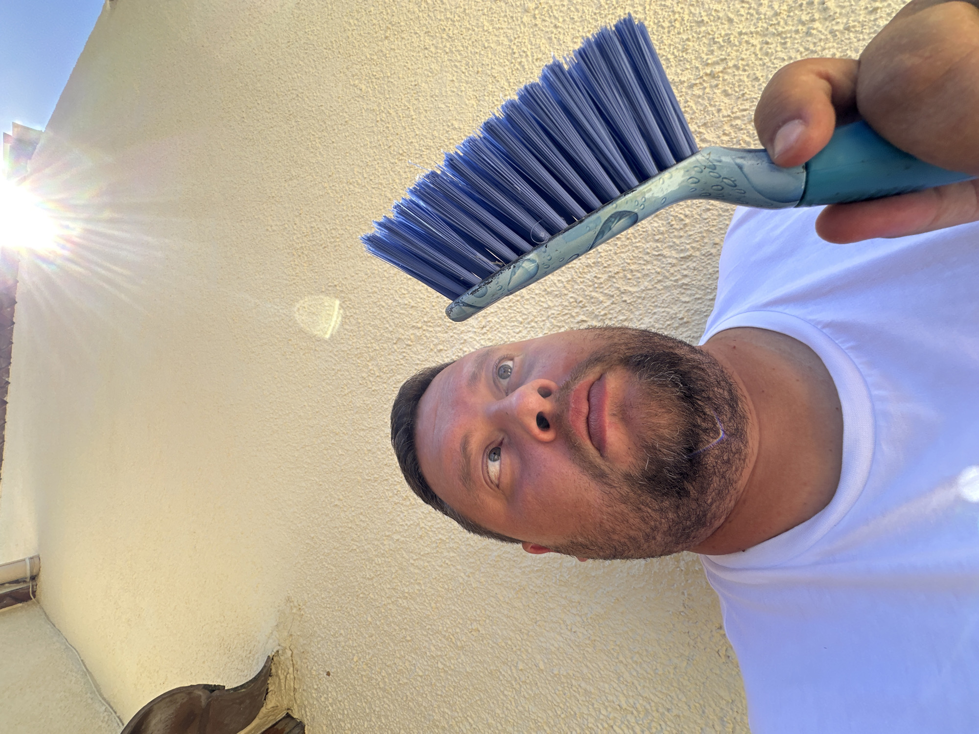A man with a beard and a white t-shirt is holding a blue-handled scrub brush up towards a textured, light-colored wall. The sun is shining brightly from the top right of the frame, creating lens flare. The man is looking up and to the right, towards the sun. The scene appears to be outdoors, possibly in a residential area, given the architecture of the building. The background consists of the building's wall and a sliver of blue sky. The location is Sant Feliu de Guíxols, Spain.