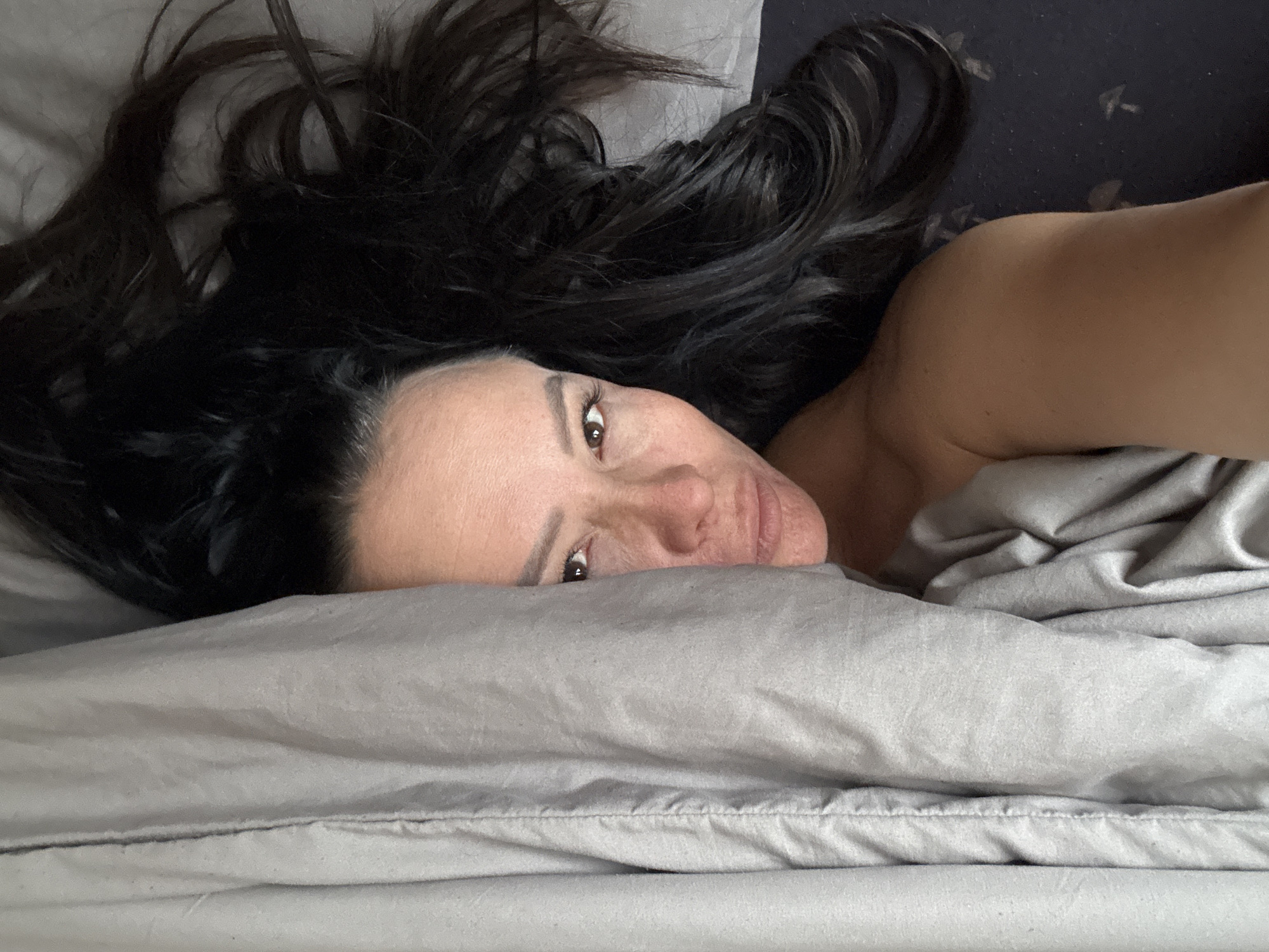 The image captures a woman lying in a bed, her head resting on a light grey pillow, and her body partially covered by matching light grey bedsheets. Her long, dark hair is spread out around her head and flows upwards across the pillow. Her face is turned slightly towards the viewer, with her right eye clearly visible and gazing directly at the camera. Her expression is neutral, possibly contemplative or serene.
Her bare right shoulder and upper arm are visible in the upper right portion of the frame, suggesting she is lying on her side. A darker fabric, possibly a contrasting bed covering or headboard, is visible behind her arm and parts of her hair. The lighting is soft and natural, indicating an indoor setting, likely a bedroom, possibly in the morning or a softly lit environment. No specific time of day, weather conditions, or any text are discernable from the image itself. The scene is set in Chaikovsky, Russia.