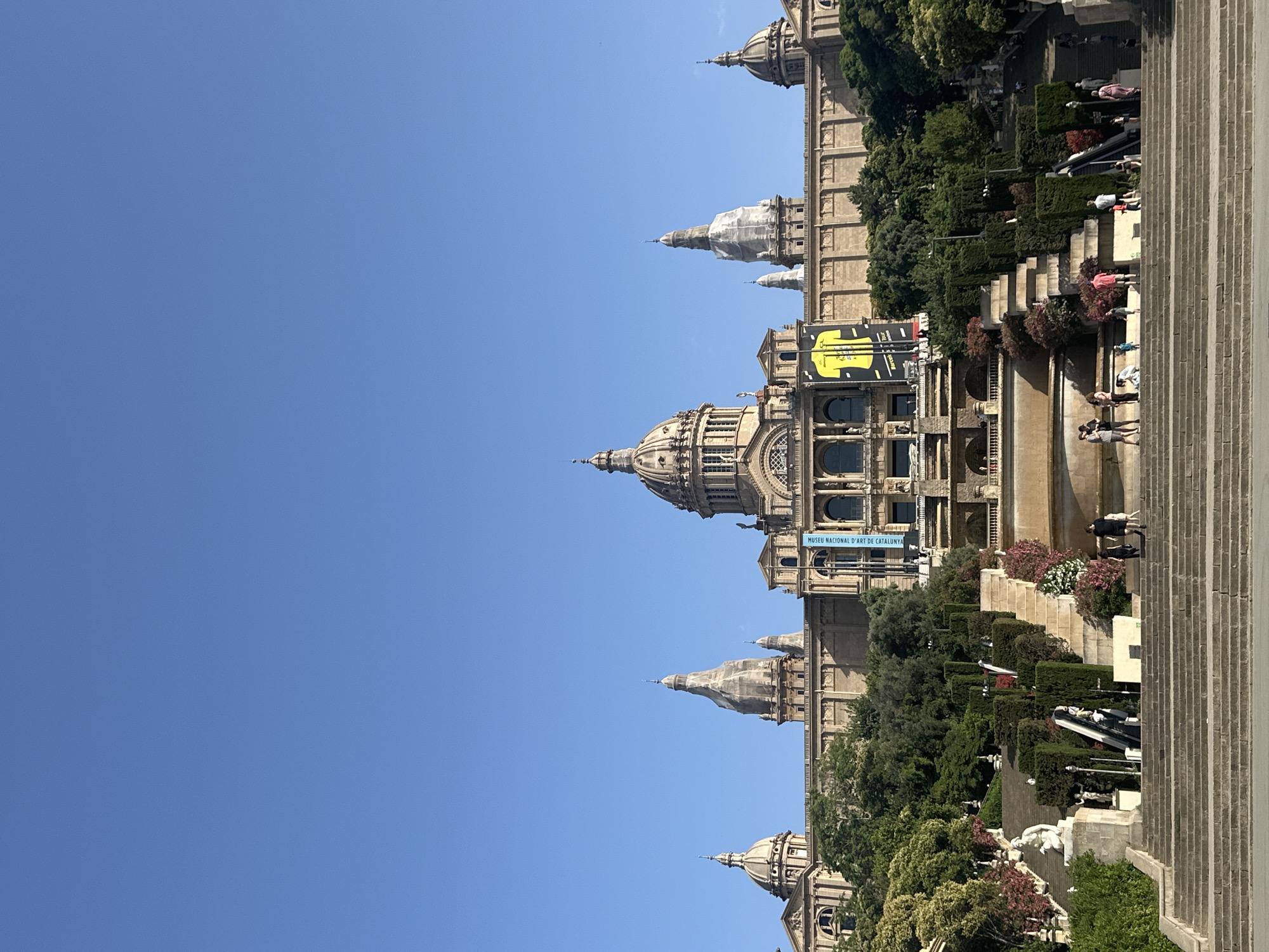 The image captures a wide-angle view of the Palau Nacional, a national palace in Barcelona, Spain, situated on Montjuïc hill. The imposing structure, built in the neoclassical style, features a prominent central dome, flanked by smaller domes and towers, all against a clear blue sky. In front of the palace, a grand cascading staircase leads up to the building, adorned with terraced gardens, sculptures, and fountains. Several people are seen ascending and descending the stairs, engaging in various activities such as walking and posing for pictures. Lush green trees and colorful flowering bushes are interspersed throughout the terraces, adding a vibrant touch to the architectural grandeur. Two banners are visible hanging from the palace façade, one displaying a yellow graphic and the other text reading "MUSEU NACIONAL D'ART DE CATALUNYA." The overall scene conveys a sense of historical significance and public gathering, with the architectural marvel and its surrounding landscape forming a picturesque backdrop.