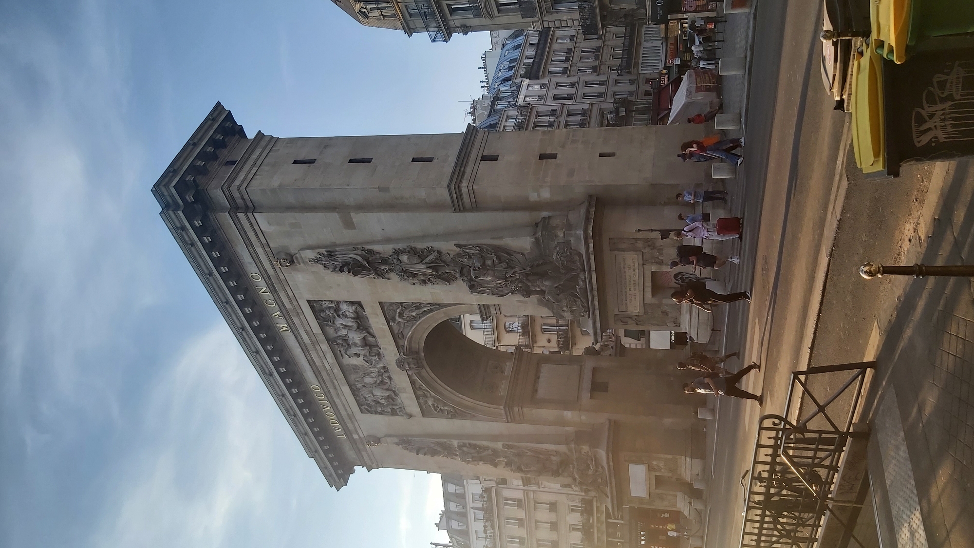 The image captures the Arc de Triomphe du Carrousel in Paris, France, under a bright, partly cloudy sky. The triumphal arch, constructed of stone, dominates the frame. Golden lettering on the arch reads "LVDOWICO MAGNO." Intricate relief sculptures adorn the arch's facade, depicting historical scenes and figures. The arch is situated in a public space with a street in the foreground and several multi-story buildings lining the background, characteristic of Parisian architecture.
In the middle ground, pedestrians are crossing the street near a crosswalk. A man in a grey t-shirt and dark pants walks across the street, while others are seen walking or standing. A person with a red suitcase is visible among the group. A black metal railing with diagonal supports runs along the lower left side of the image, partially obscuring the view of the street and sidewalk. Two large trash receptacles with yellow lids are present in the lower right foreground. The lighting suggests it is either early morning or late afternoon, casting long shadows.