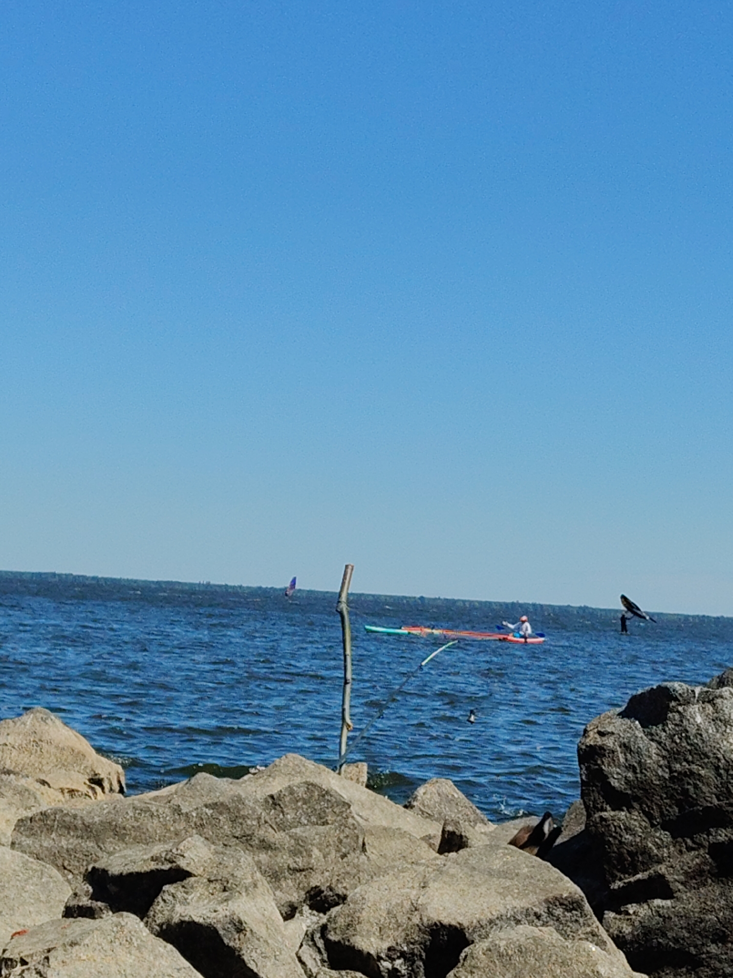 The image depicts a scene on a body of water, likely a lake or river, under a clear blue sky. In the foreground, large grey rocks form a barrier along the water's edge. The water is choppy with visible waves.
Further out on the water, there are several individuals engaged in water activities. On the left, a person is windsurfing with a blue sail visible. To the right of the windsurfer, a person is paddling a kayak or a stand-up paddleboard (SUP). The SUP appears to be orange and red. Another individual is visible in the water further to the right, appearing to be on a different type of watercraft, possibly a foilboard, with a dark wing-like structure behind them. A thin wooden pole stands upright in the water between the foreground rocks and the distant activities.
The location is identified as Cherkasy, Ukraine, suggesting this is a view of the Dnieper River or the Kremenchuk Reservoir. The overall atmosphere is bright and sunny, indicative of a pleasant day for outdoor recreation.