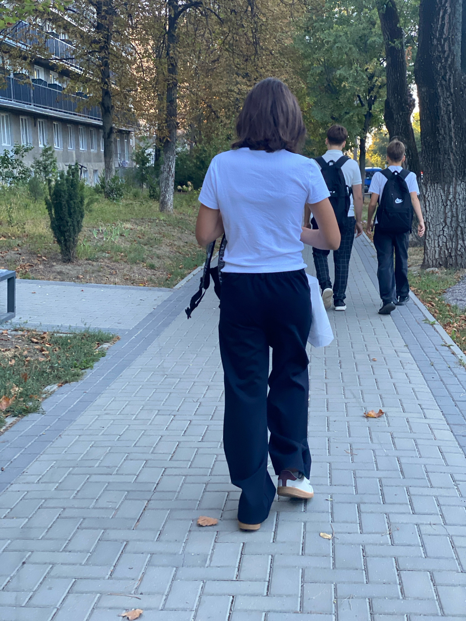 The image shows three individuals walking away from the camera on a brick sidewalk. The person in the foreground is wearing a white t-shirt and black pants, carrying a white bag. Behind them, two other individuals, presumably teenagers, are also walking, both wearing t-shirts and backpacks. The one in the middle wears a white t-shirt and patterned pants, while the one on the right wears a white t-shirt and black pants. The environment appears to be an urban setting with trees and a building visible in the background. There are fallen leaves on the ground, suggesting it is autumn. The building in the background is a multi-story structure with balconies and windows, characteristic of residential buildings in Chișinău, Moldova. The sidewalk is paved with gray rectangular bricks arranged in a herringbone pattern. The lighting suggests it is daytime, possibly late afternoon, with sunlight filtering through the trees.