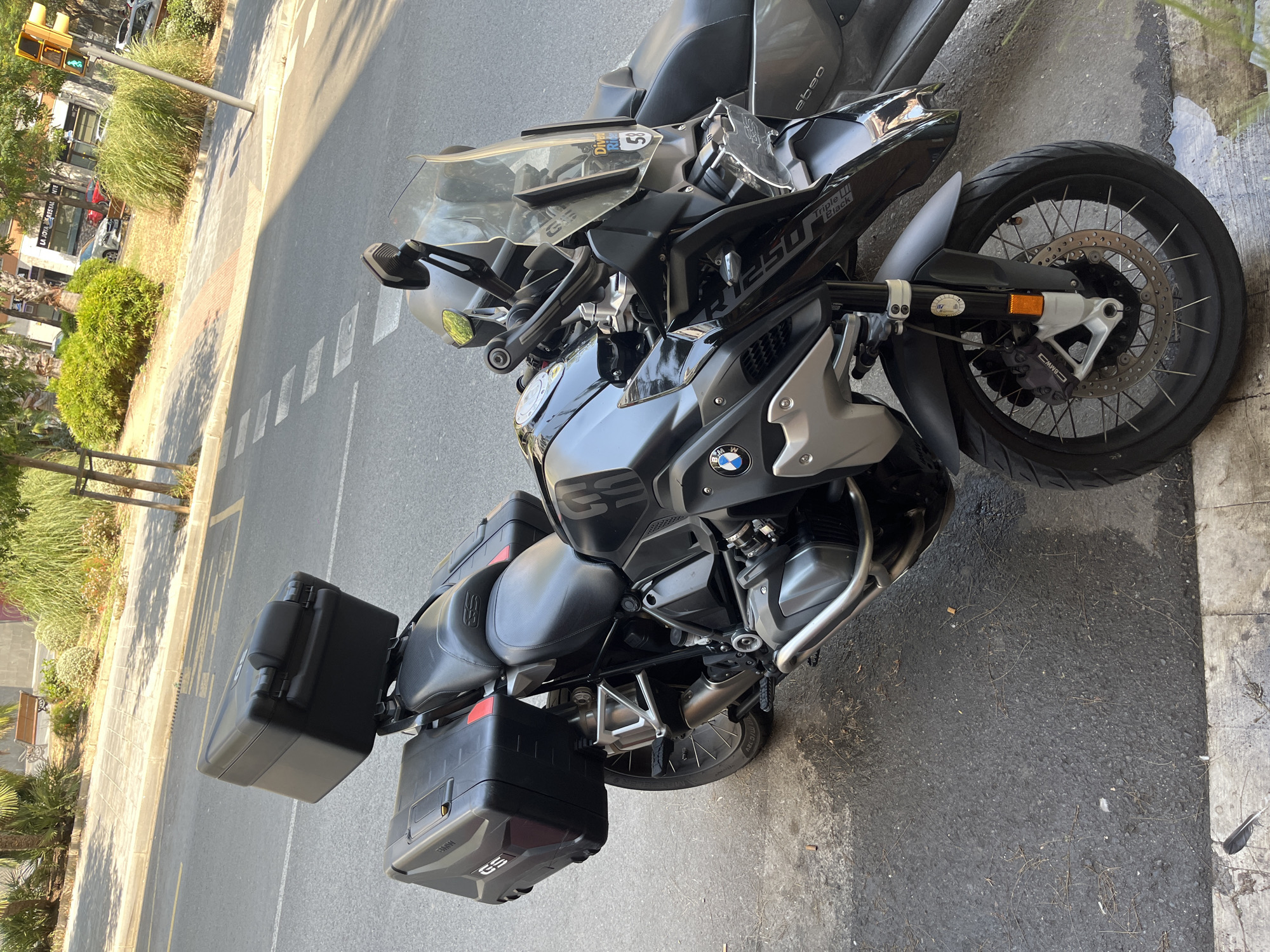 A black BMW R1250GS motorcycle is parked on a city street in Tarragona, Spain, on a sunny day. The motorcycle is equipped with large black panniers on both sides and a top case. The street has a pedestrian sidewalk with some greenery and trees. In the background, there are buildings and a traffic light. The motorcycle is facing forward, slightly angled to the left. The scene is static, with no people or activity evident.